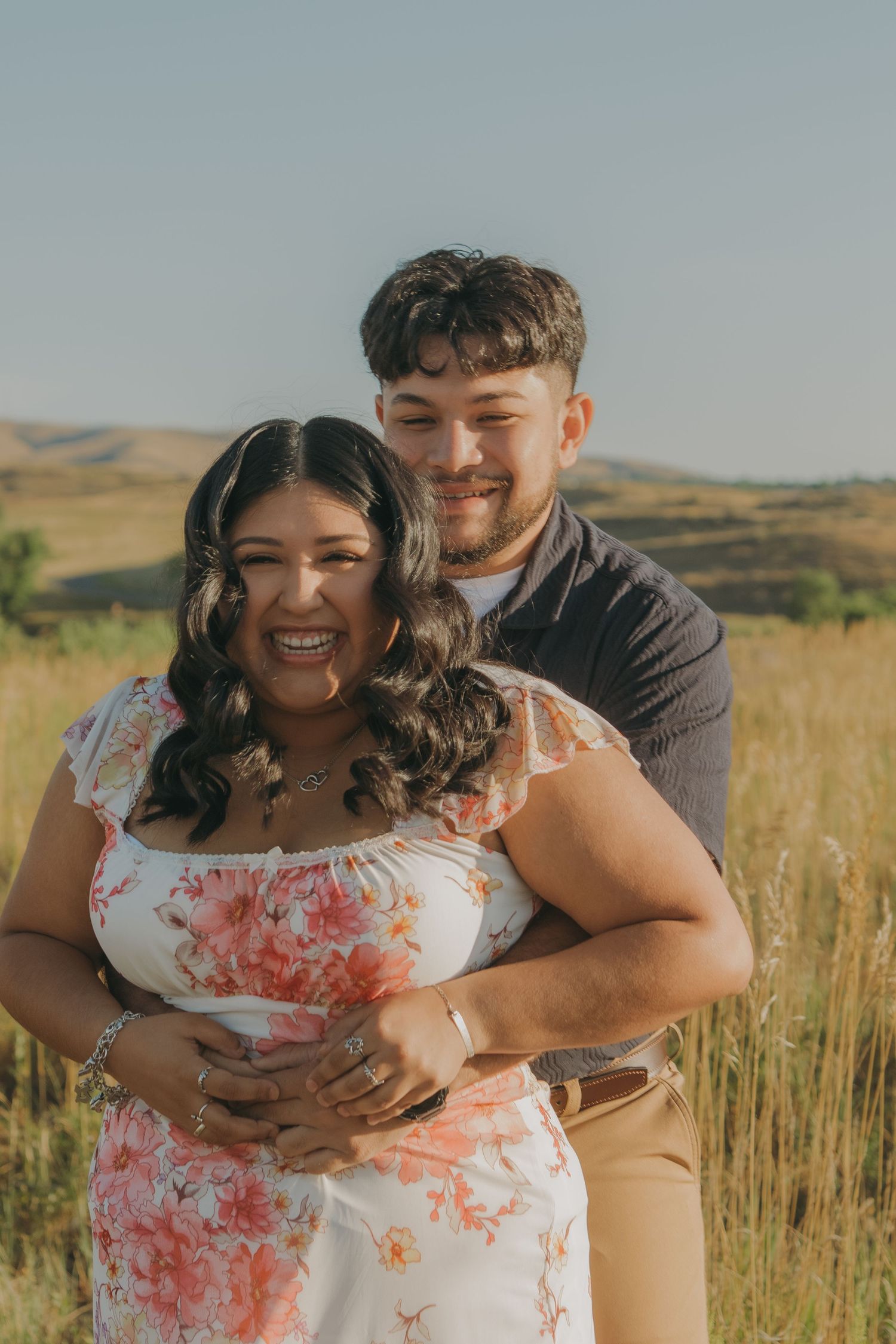 A joyful couple laughs together in golden afternoon light amidst tall grass and hills.