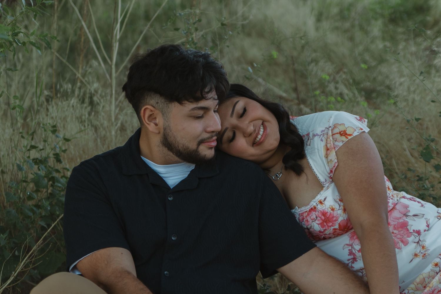 A couple shares a tender moment while sitting together in tall grass during golden hour.