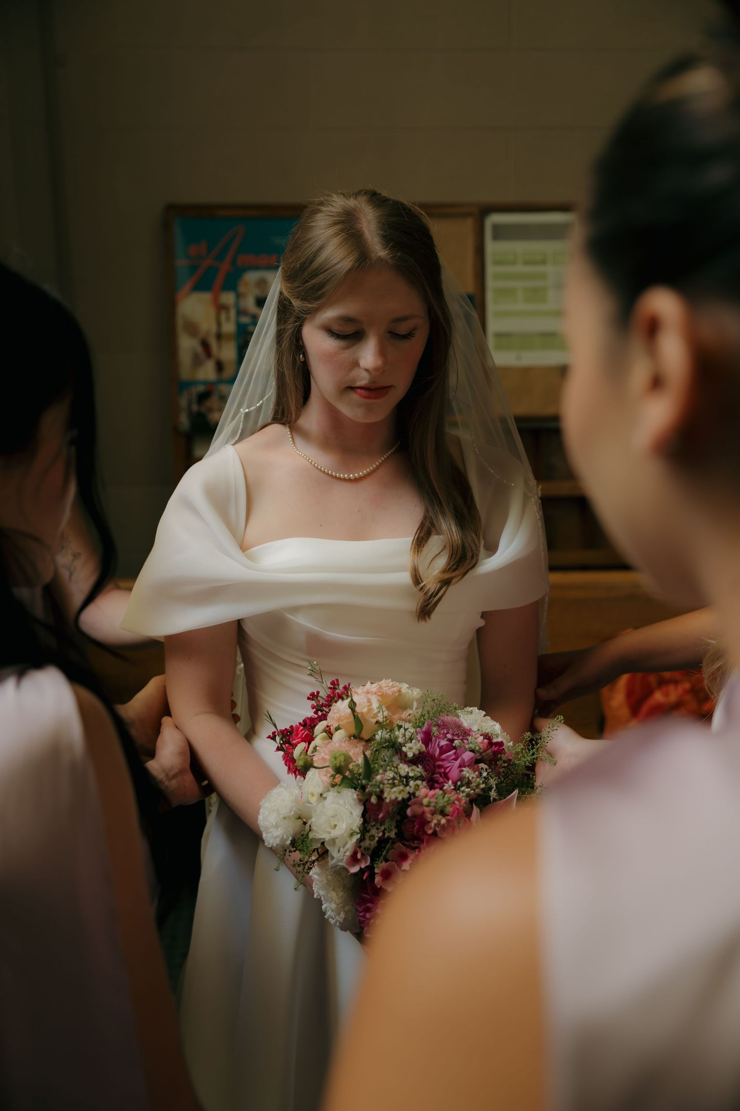 Bride in white off-shoulder gown holds colorful bouquet while bowing head in prayer with bridesmaids before wedding ceremony.