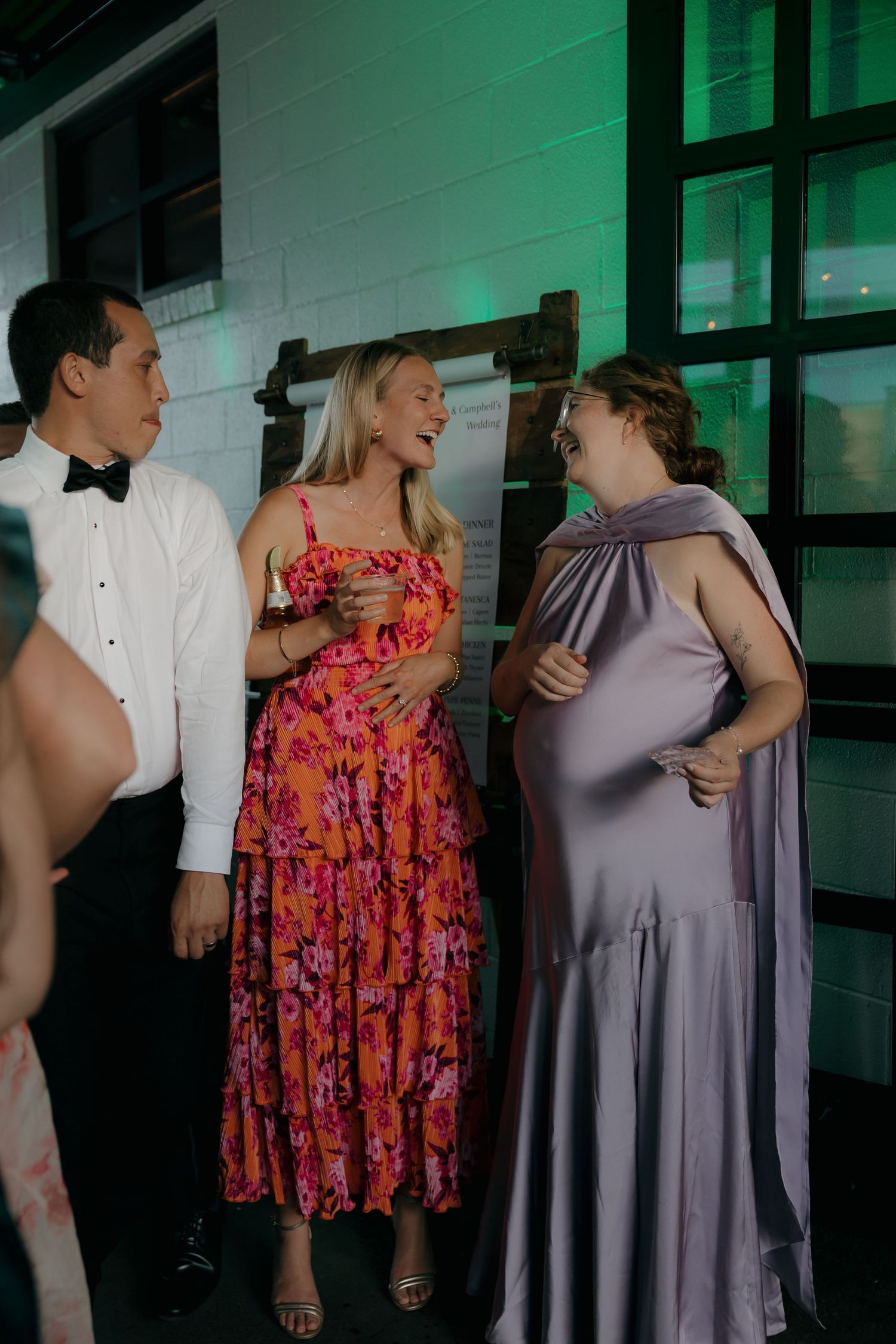 Wedding guests in formal attire laughing together near green-lit white brick wall at reception.