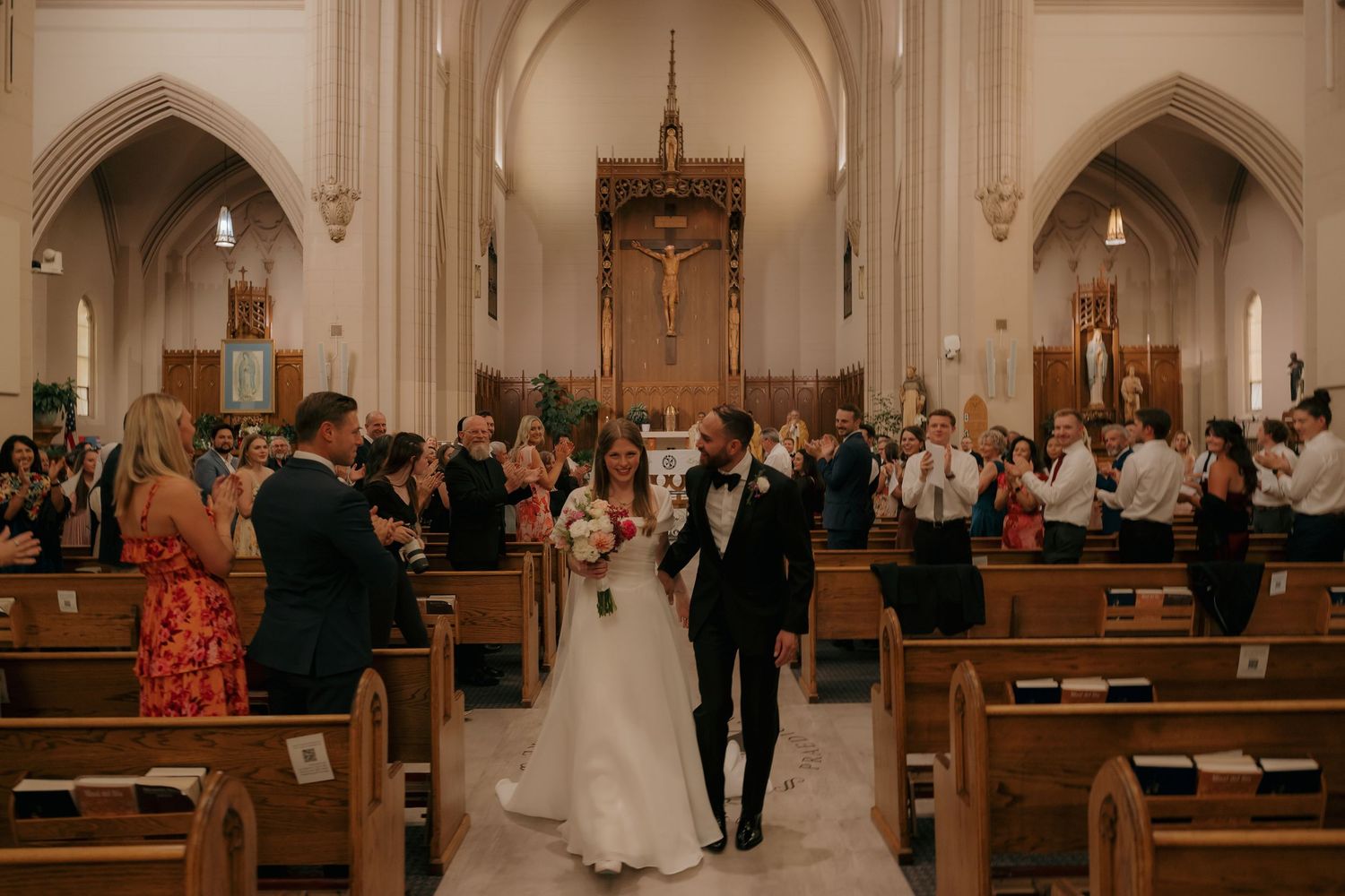 Bride and groom walk down church aisle after wedding ceremony, surrounded by cheering guests in elegant sanctuary.