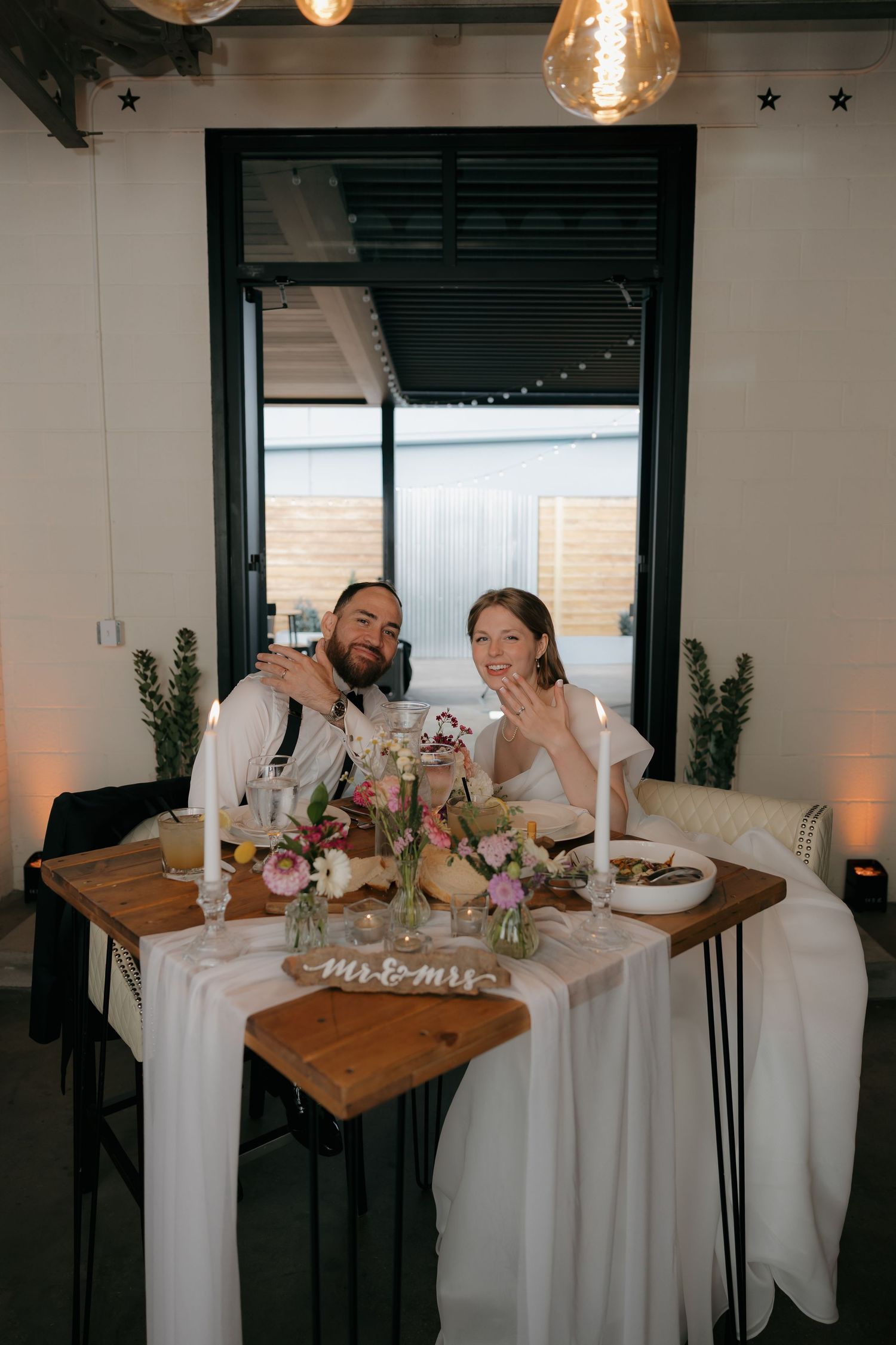 Newlyweds smile and show rings at sweetheart table decorated with colorful flowers and Mr and Mrs sign.