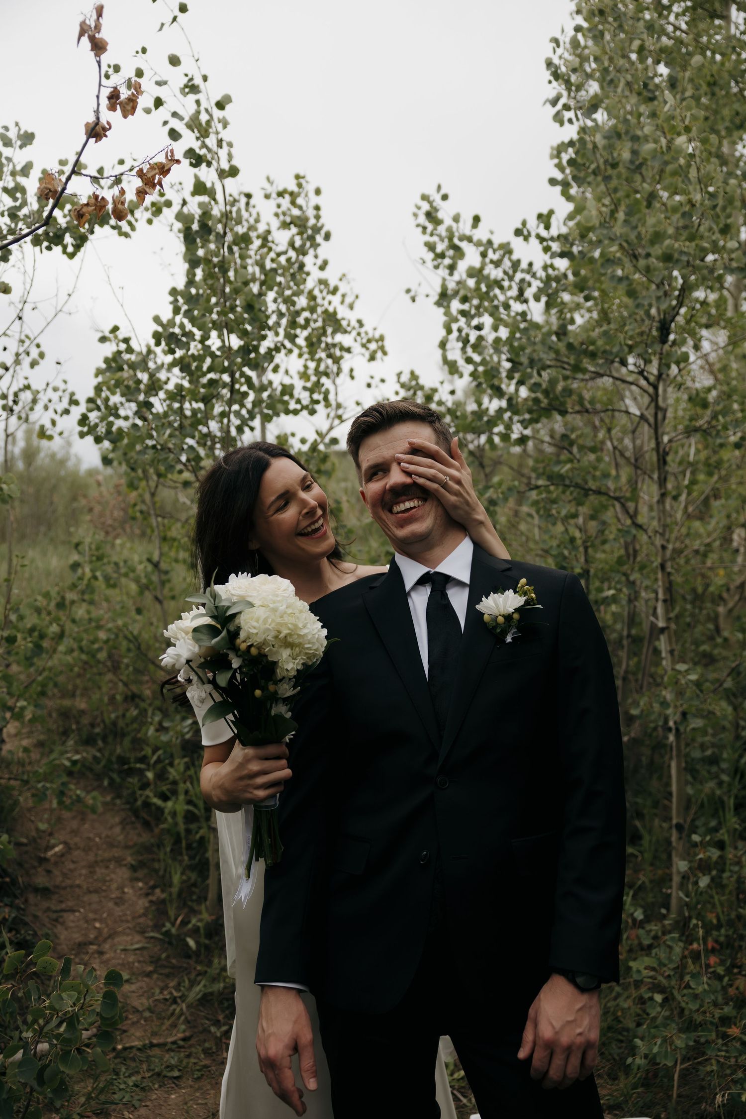 Laughing bride covers grooms eyes with her hand during playful outdoor wedding photo in forest.