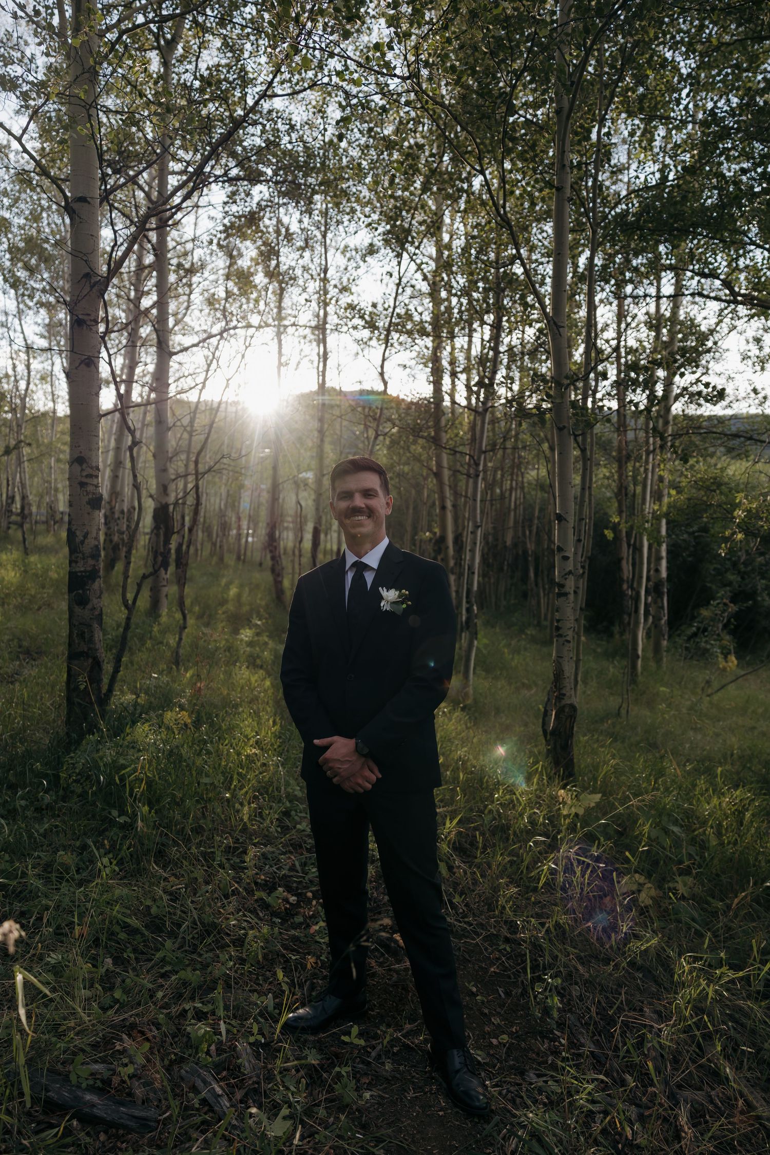 Smiling groom in black suit stands among birch trees with sunlight streaming through forest.