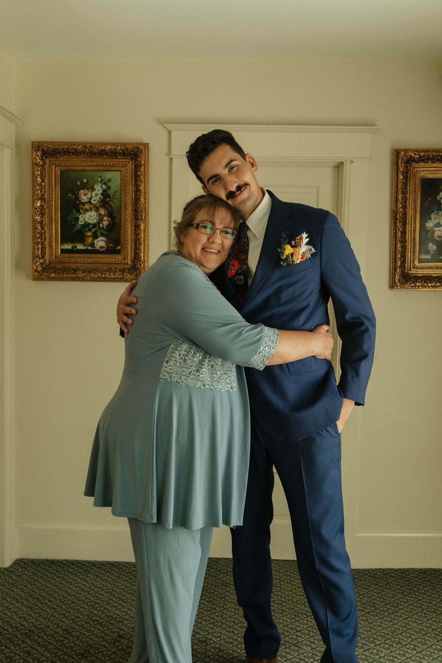 Heartwarming moment between loved ones during wedding preparations in a room with framed artwork.