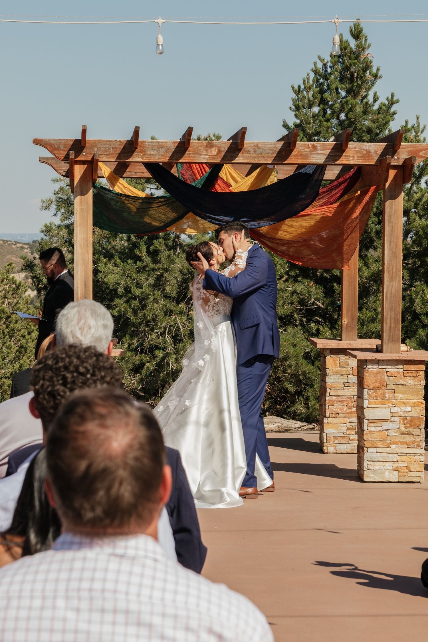 A wedding ceremony takes place under a wooden pergola against a scenic mountain backdrop.