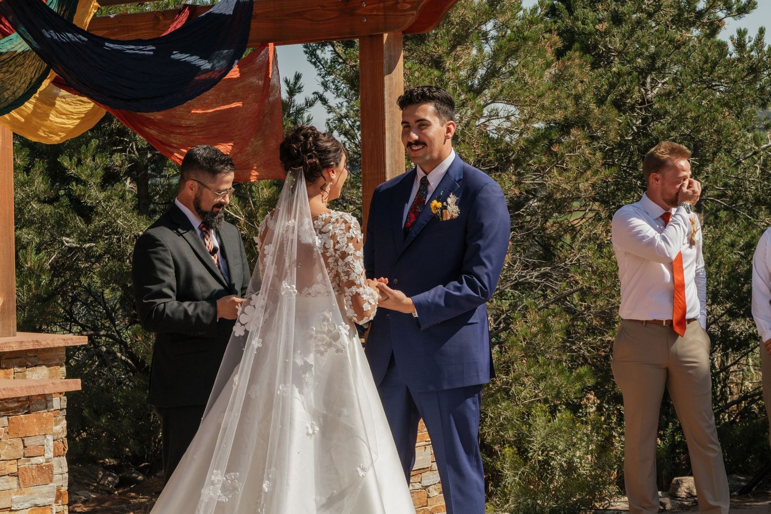Outdoor wedding ceremony under a rustic wooden pergola with gathered guests and natural scenery.