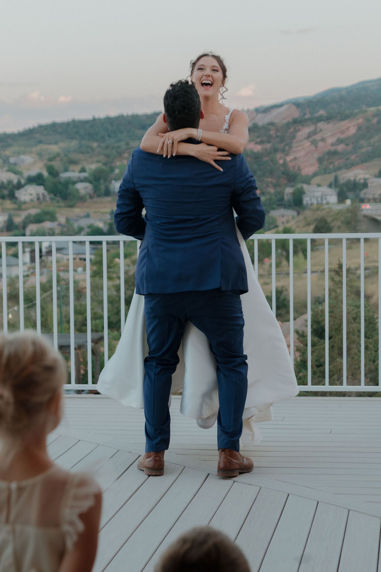 Wedding couple shares a joyful moment on a deck while guests watch from below.