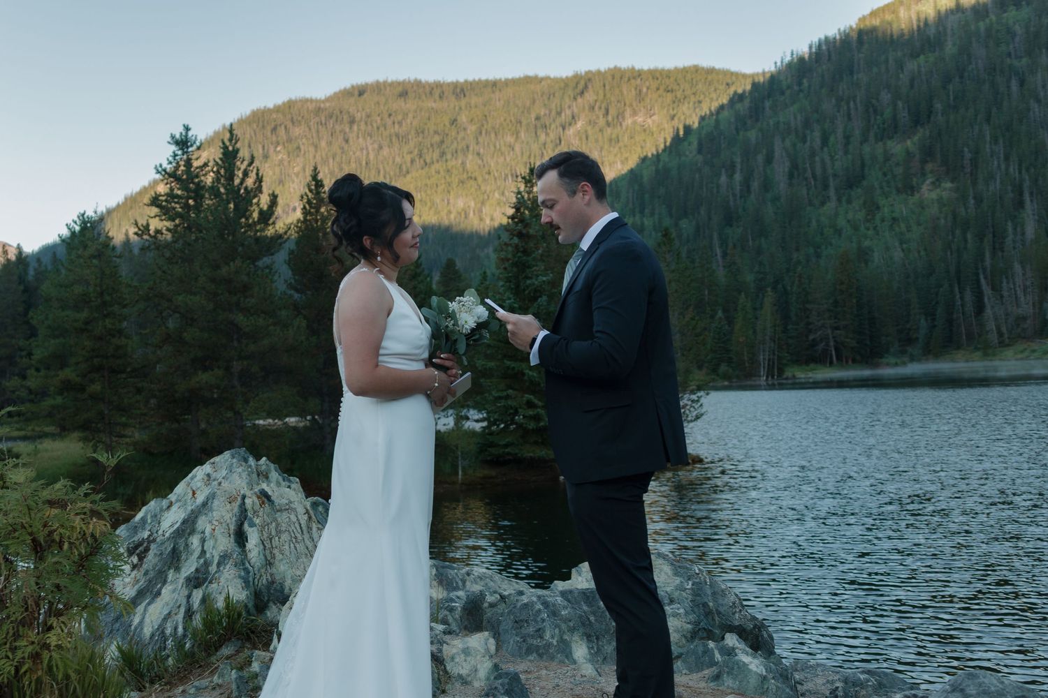 A couple shares an intimate moment at dusk beside a serene mountain lake surrounded by evergreen forests.