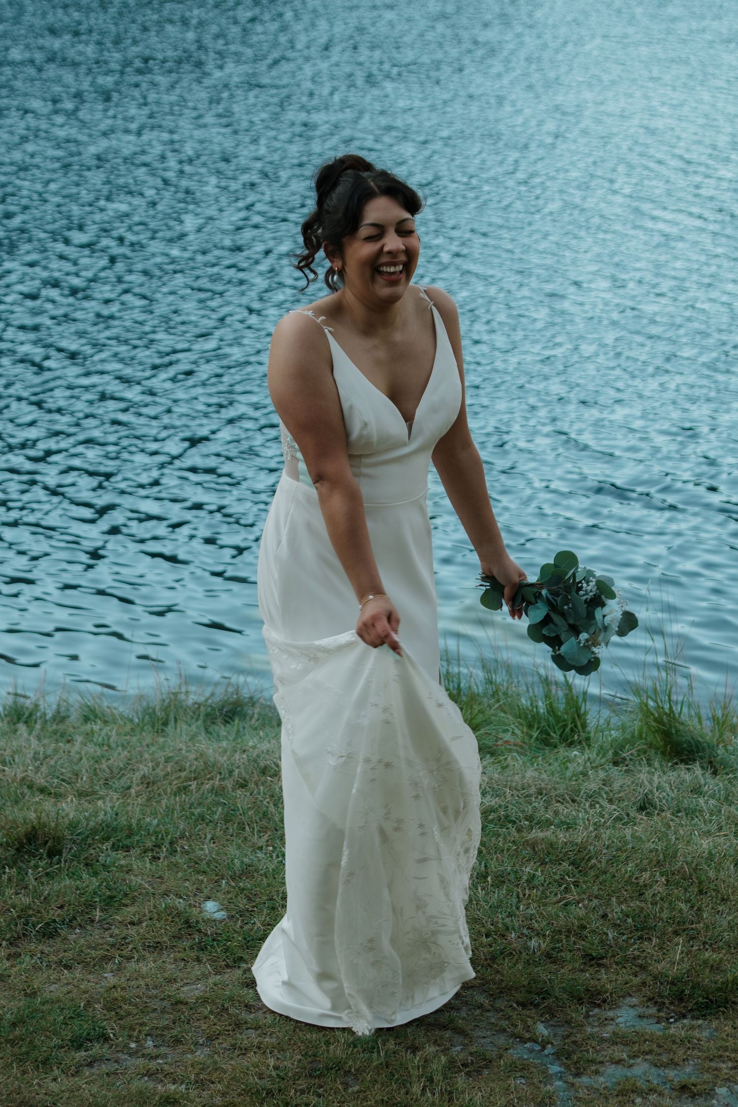 Person in white wedding dress laughing joyfully by a serene lake.