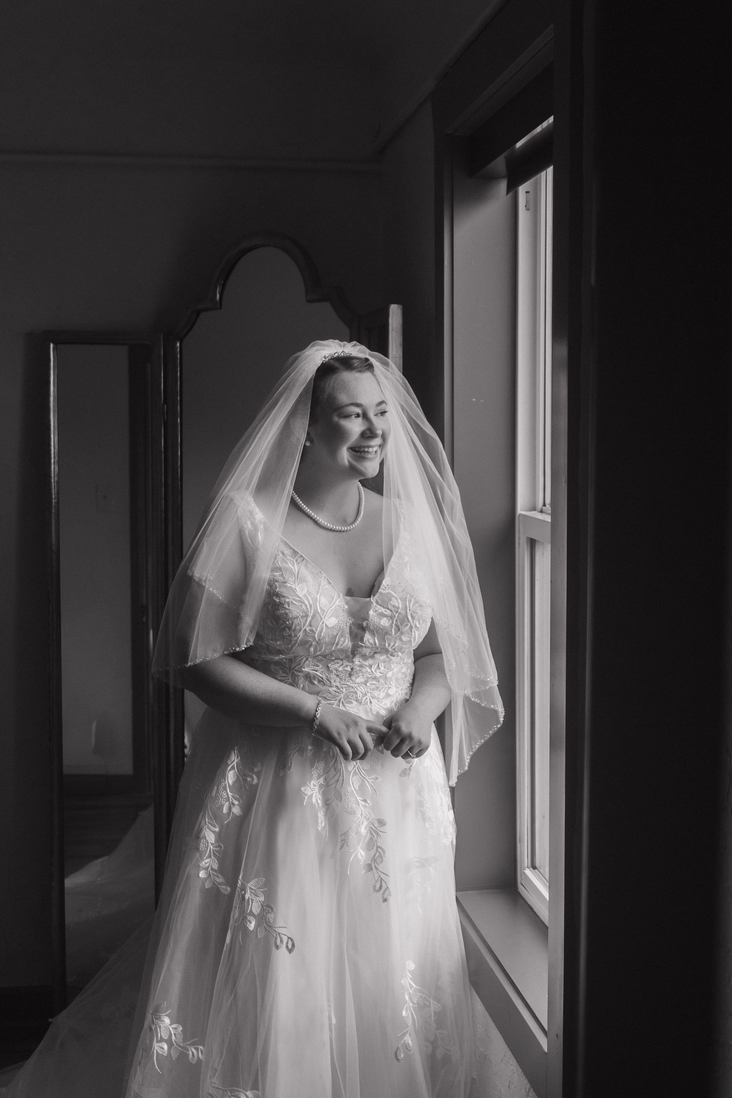 A bride in a white lace wedding dress and veil stands by a window in black and white photography.