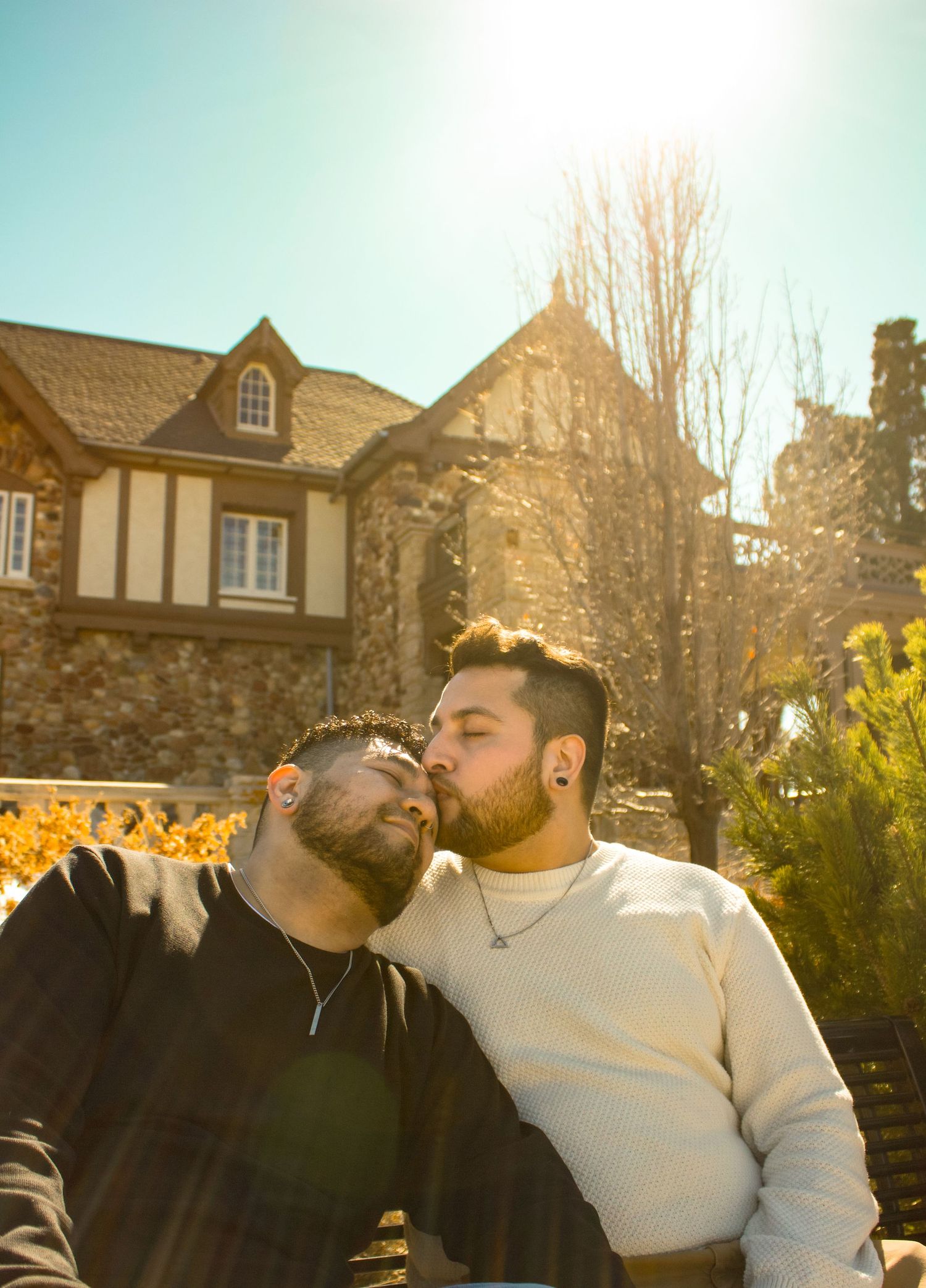 A couple sharing an intimate moment on a bench in front of a Tudor-style stone building with autumn foliage.
