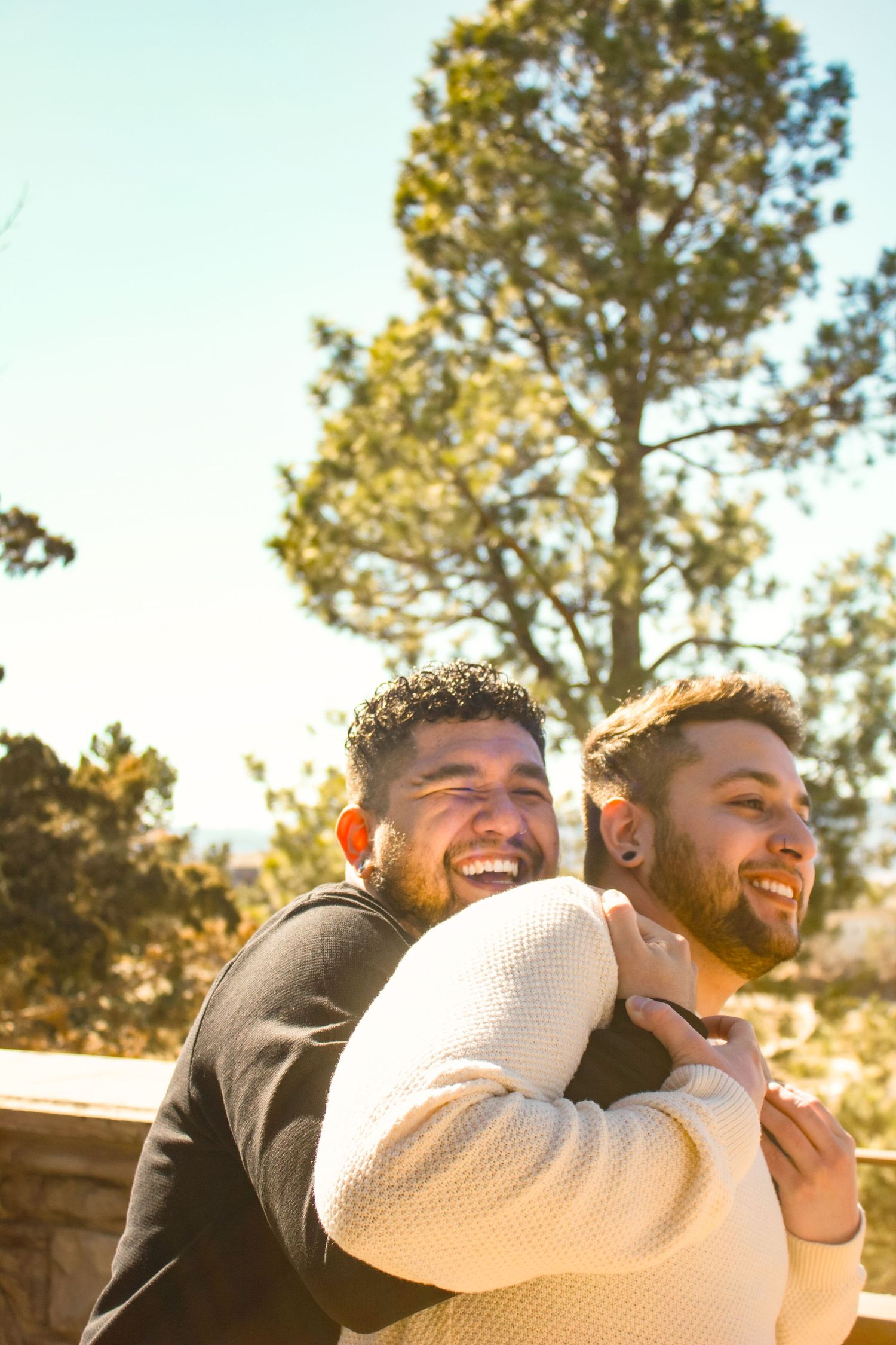 Two people embrace joyfully outdoors with sunlit pine trees in the background.