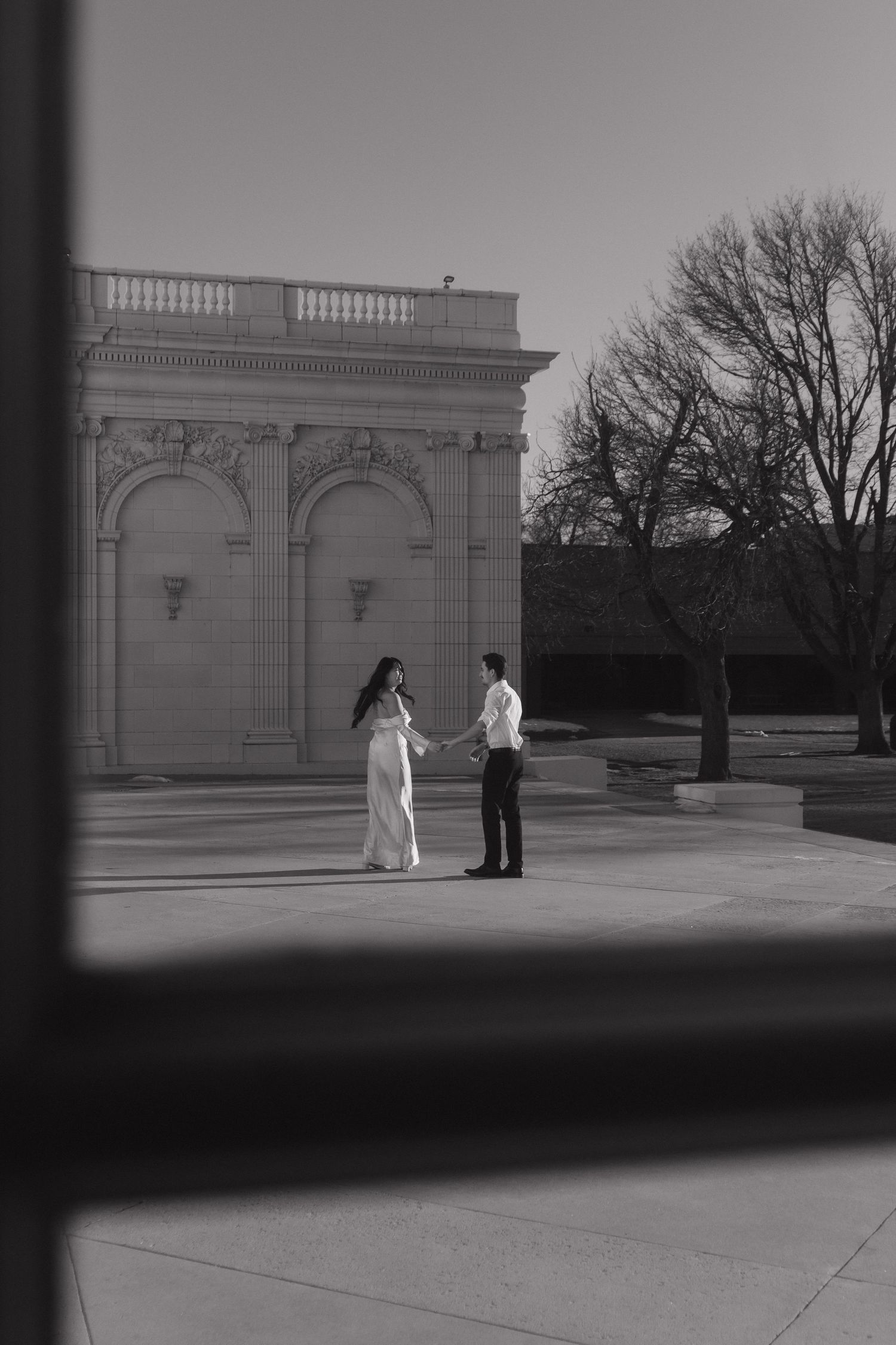 Two figures stand in silhouette against a classical architectural backdrop in black and white.