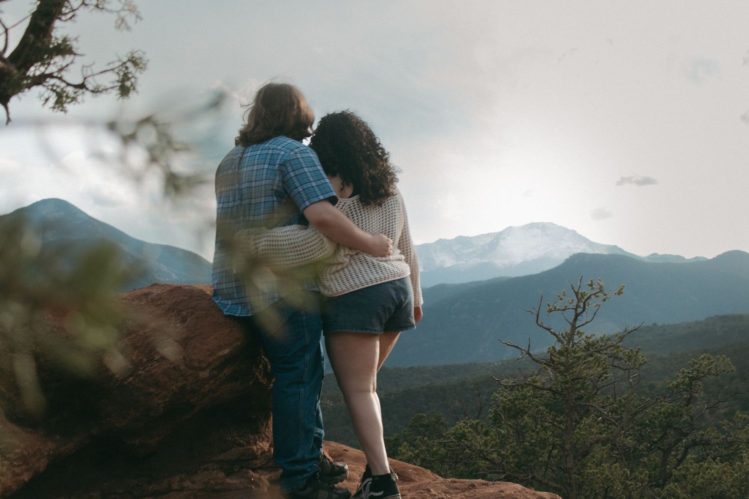 Two figures stand together on a cliff edge overlooking a mountain valley at dusk.