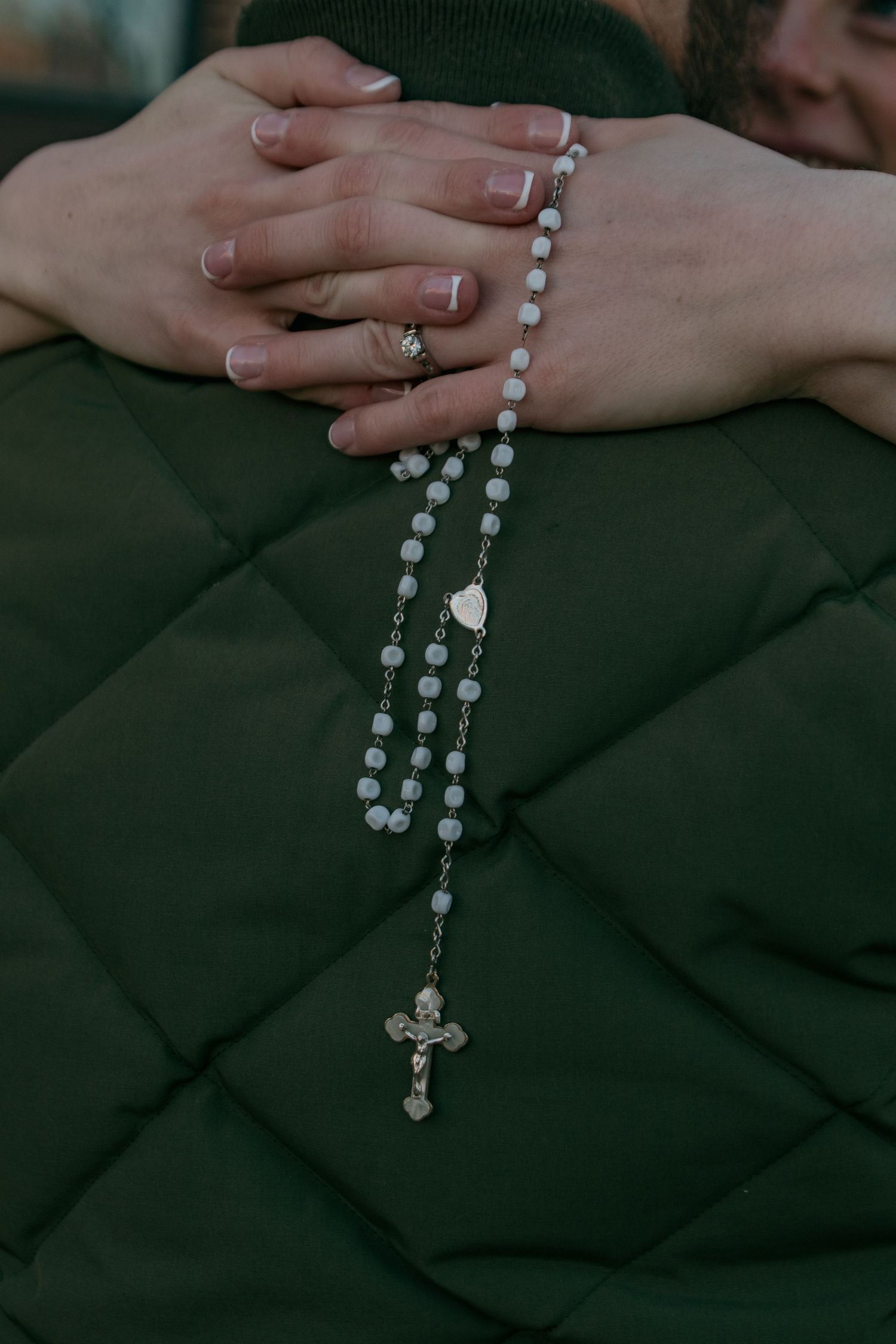Hands holding a silver rosary with white beads against a dark quilted fabric background.