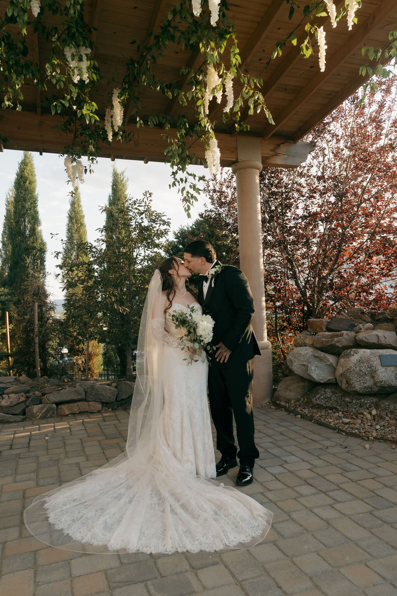 Bride and groom share a kiss under a floral pergola, bride in lace gown with veil and bouquet.