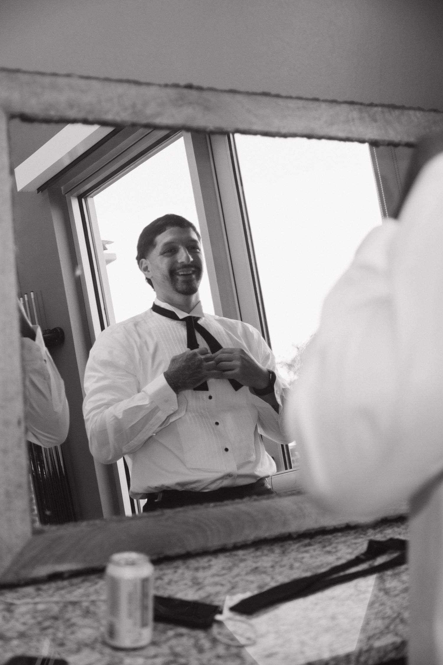 Black and white photo of groom smiling while adjusting tie in mirror before wedding ceremony.