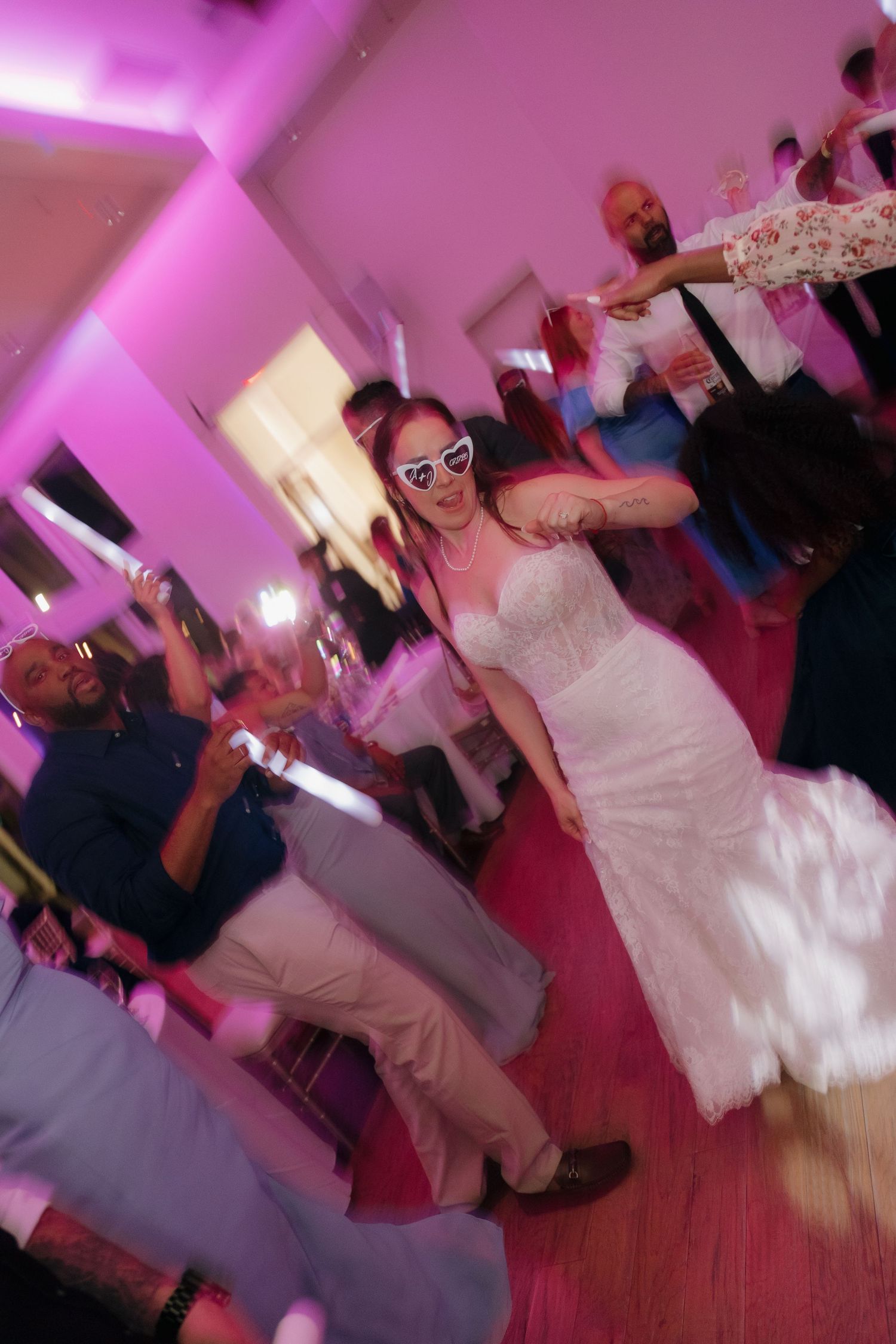 Bride wearing heart-shaped sunglasses dancing joyfully with guests at wedding reception.