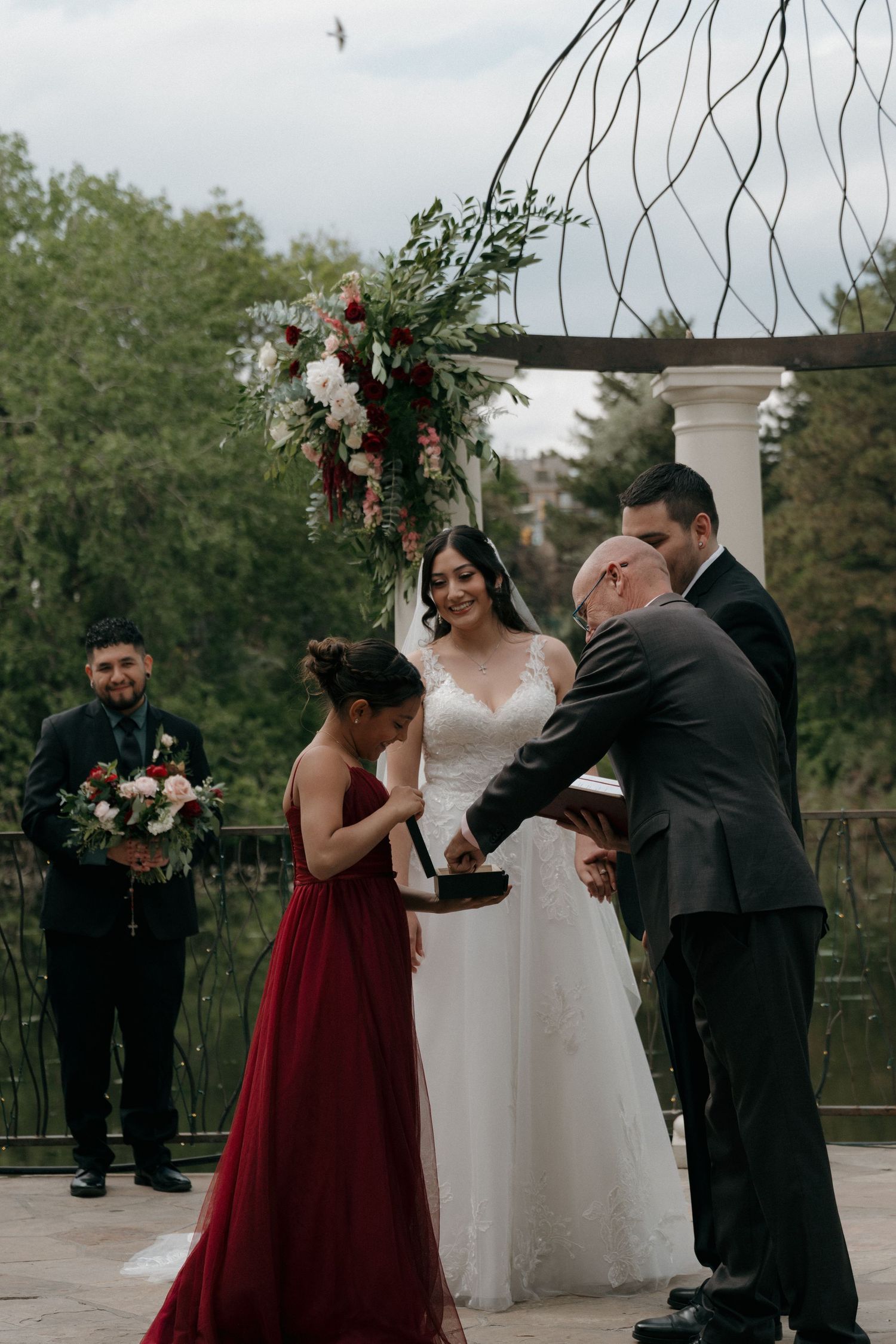 Bride and groom exchange rings at outdoor ceremony under floral arch, officiant and bridesmaid in red assist.