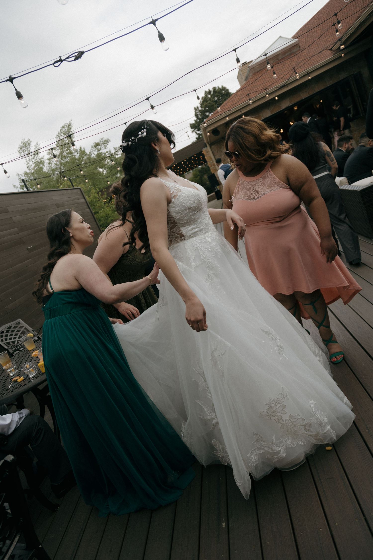 Bridesmaids in green and pink dresses assist bride in lace gown on an outdoor wedding deck.