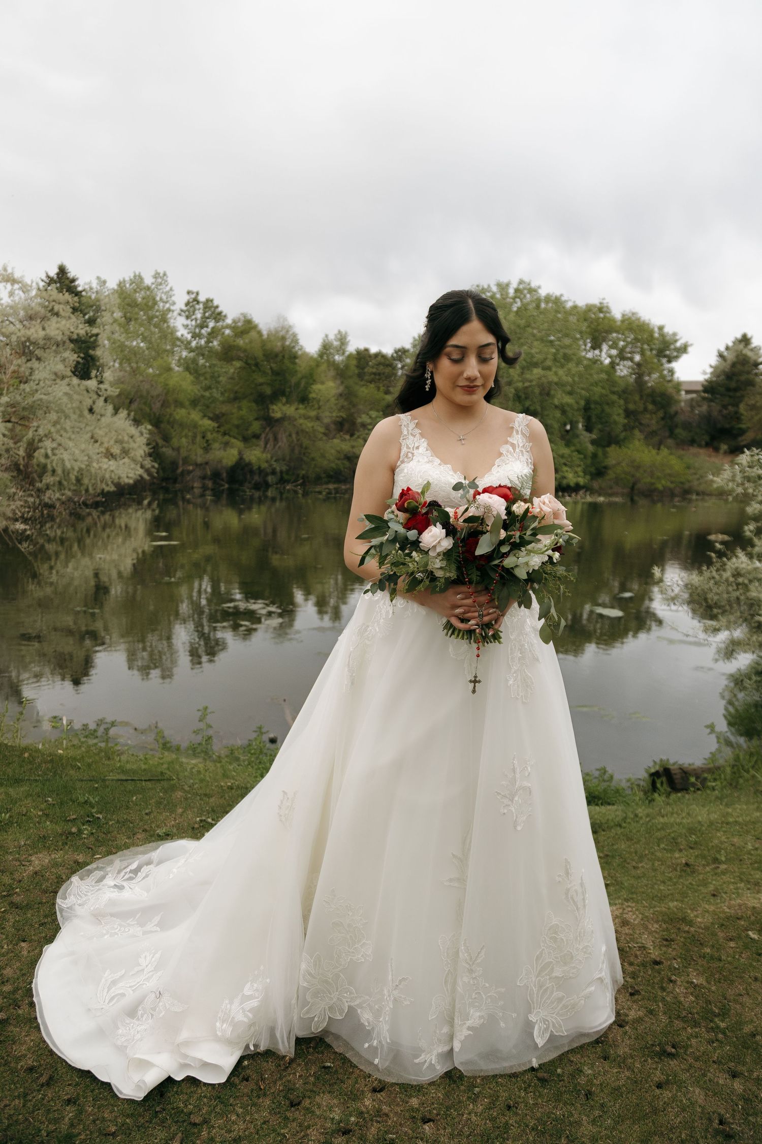Elegant bride in lace A-line gown holds red and white bouquet while standing before a serene lake on her wedding day.