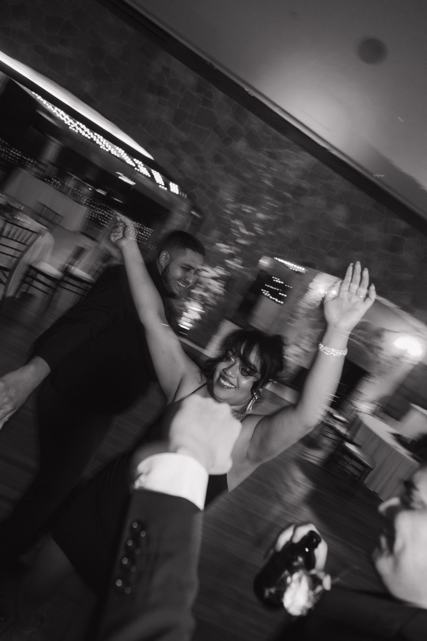 Black and white motion blur photo of smiling woman dancing with hands raised at wedding.