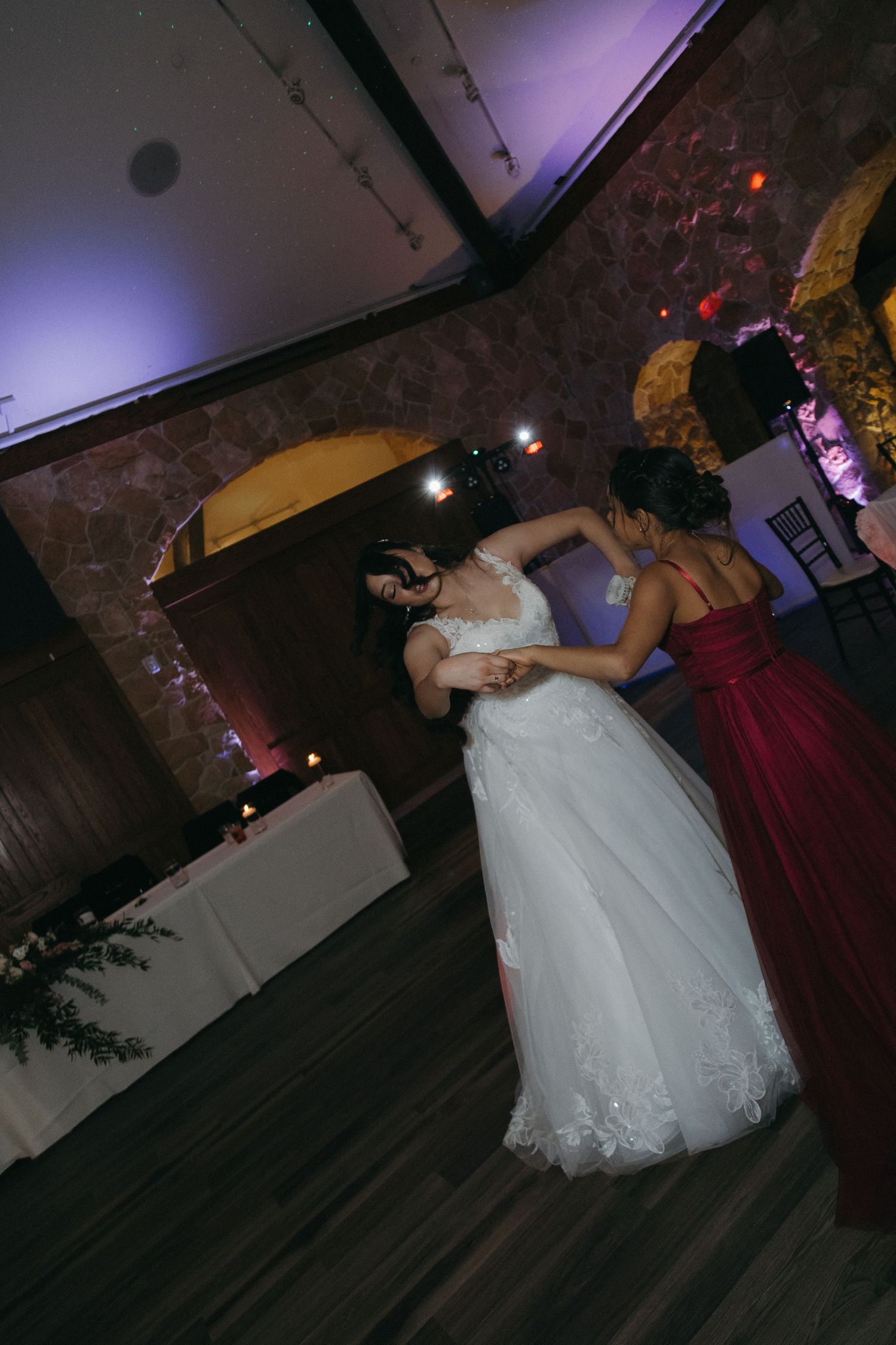 Bride in white lace gown dances with bridesmaid in red dress at wedding reception.