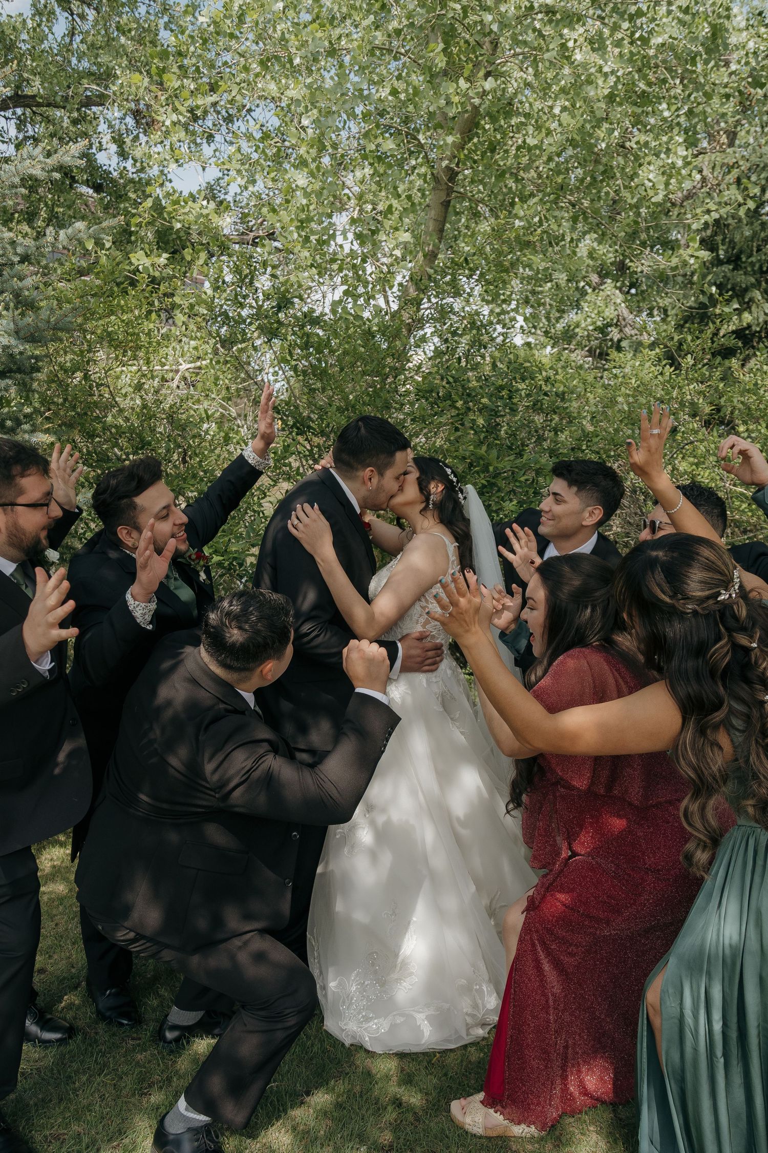 Wedding party surrounds bride and groom with cheers as couple shares a kiss outdoors.