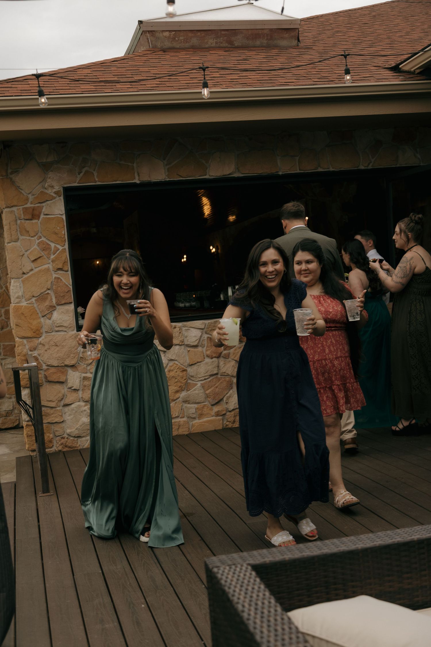 Three women in formal gowns smile and hold drinks on a wooden deck at an outdoor wedding reception.