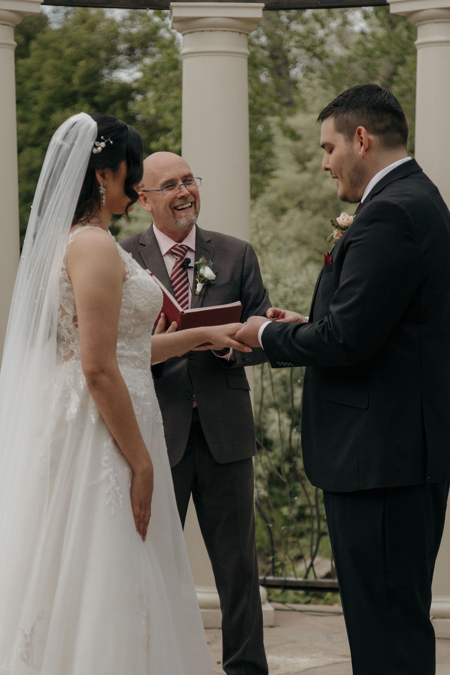 Smiling officiant watches as groom places ring on brides hand during outdoor wedding ceremony with columns.