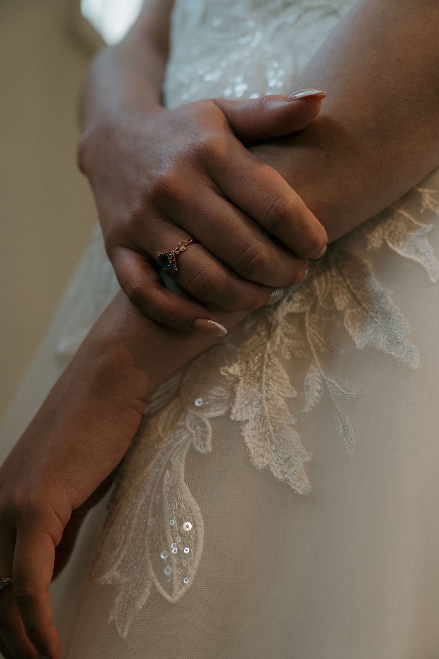 Close-up of bride's hands with diamond ring resting on intricate lace and sequin wedding dress.
