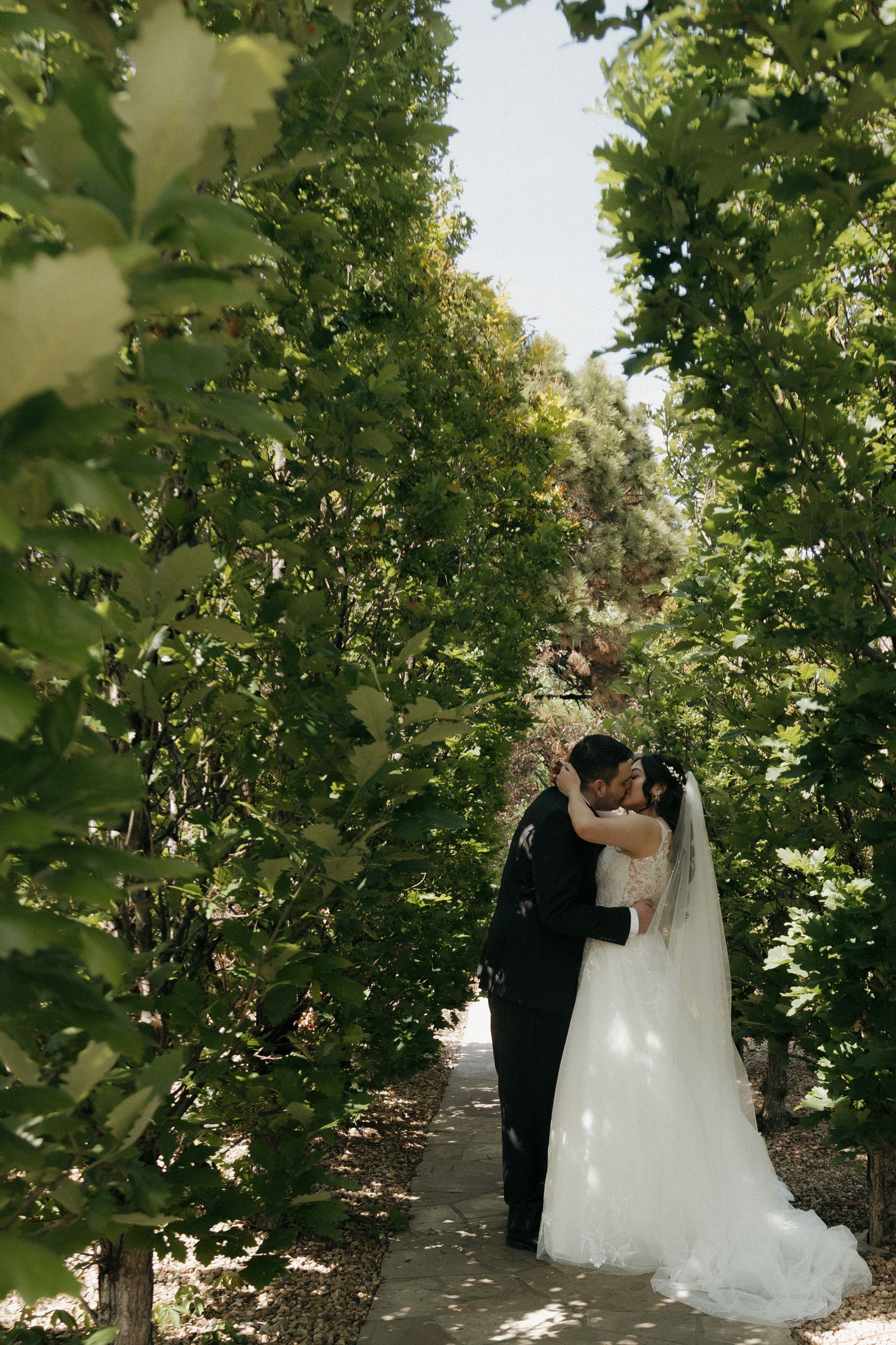 Bride and groom sharing a romantic kiss along a lush green tree-lined garden pathway on their wedding day.
