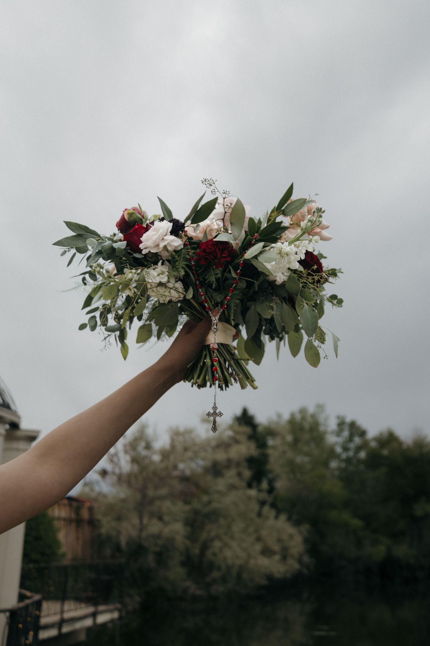 A hand holds a lush bridal bouquet of red, white, and pink flowers with greenery against a cloudy sky.