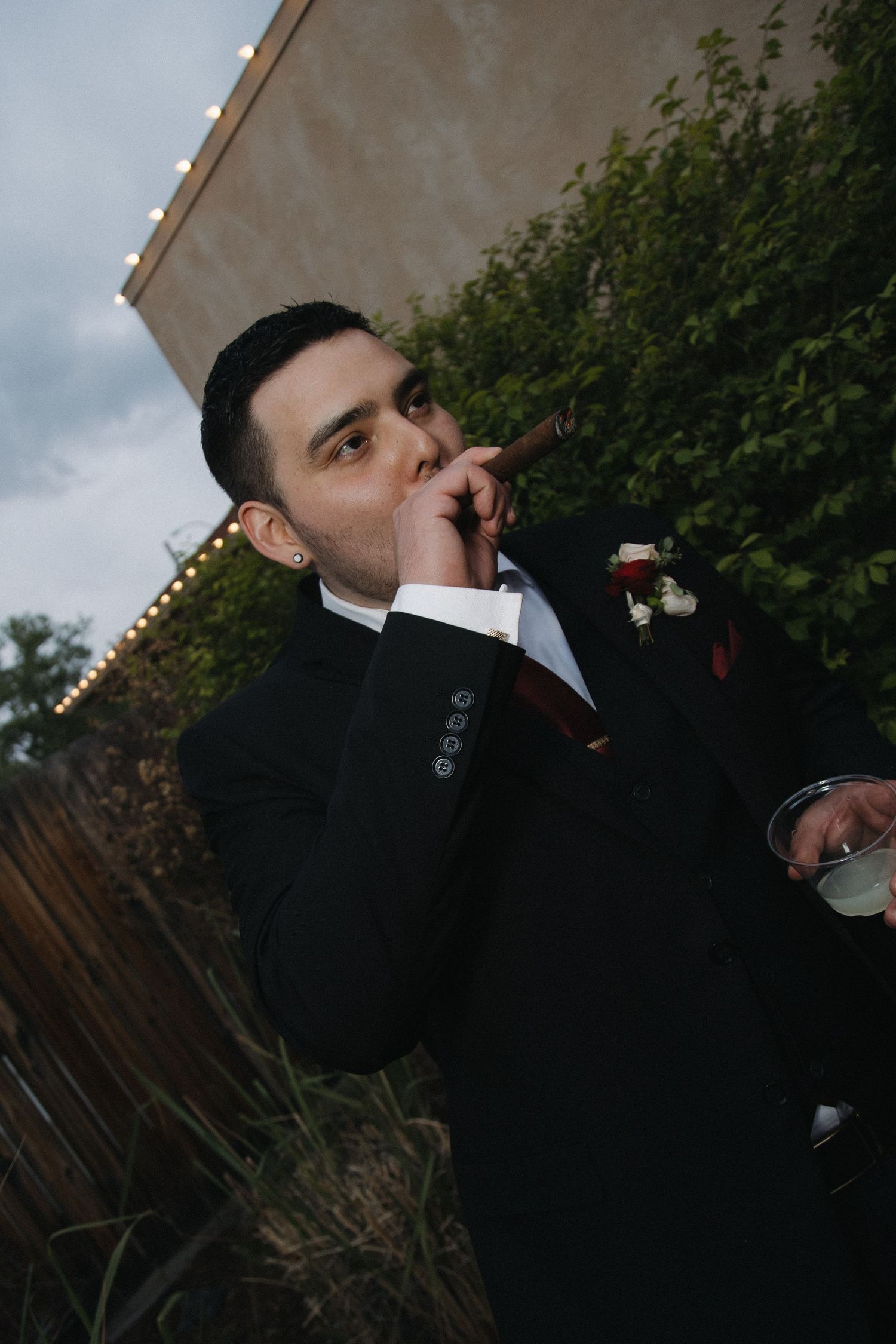 Dapper groom in black suit smoking cigar and holding drink at outdoor evening wedding reception.