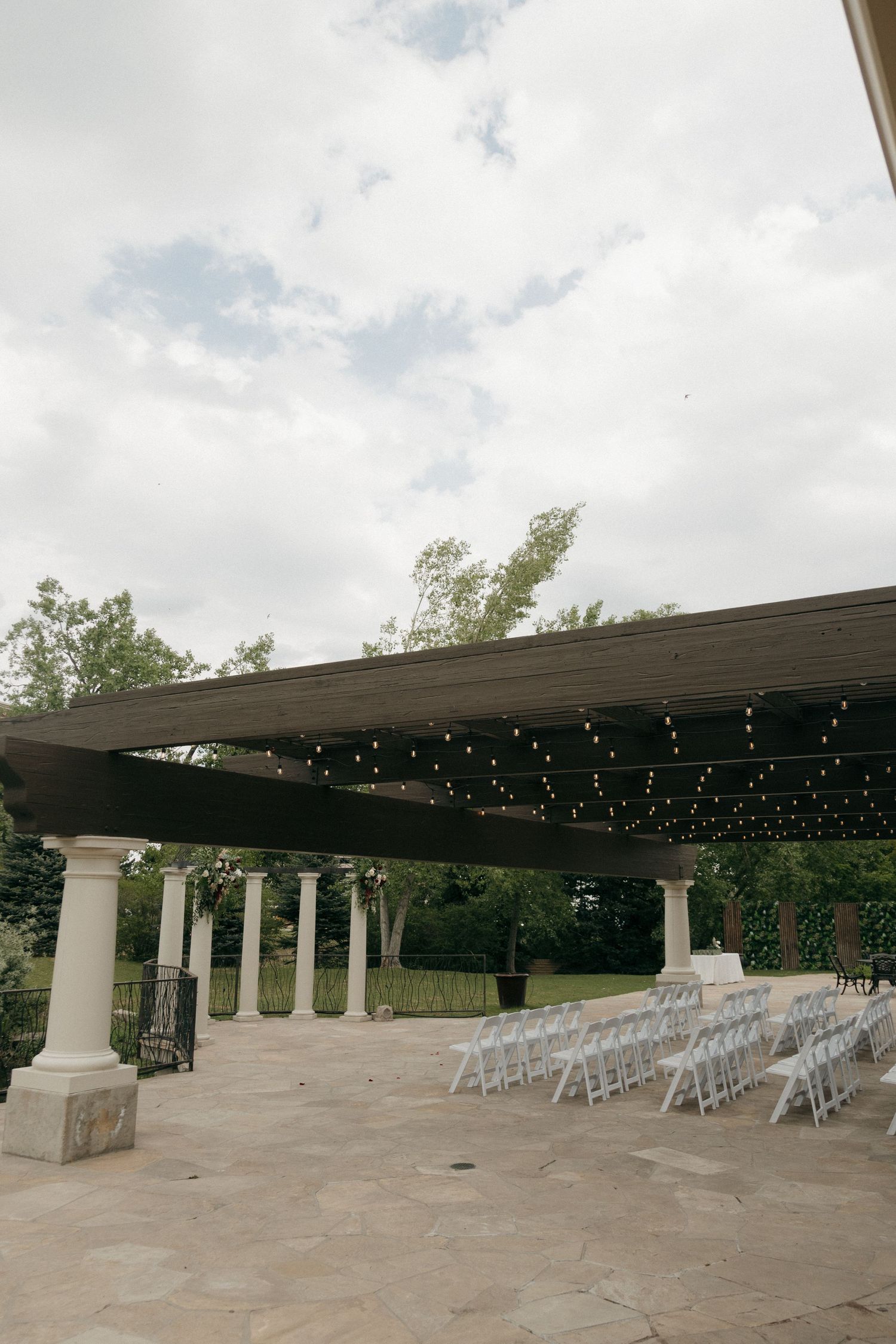 Outdoor wedding ceremony setup with white chairs under a pergola adorned with string lights.