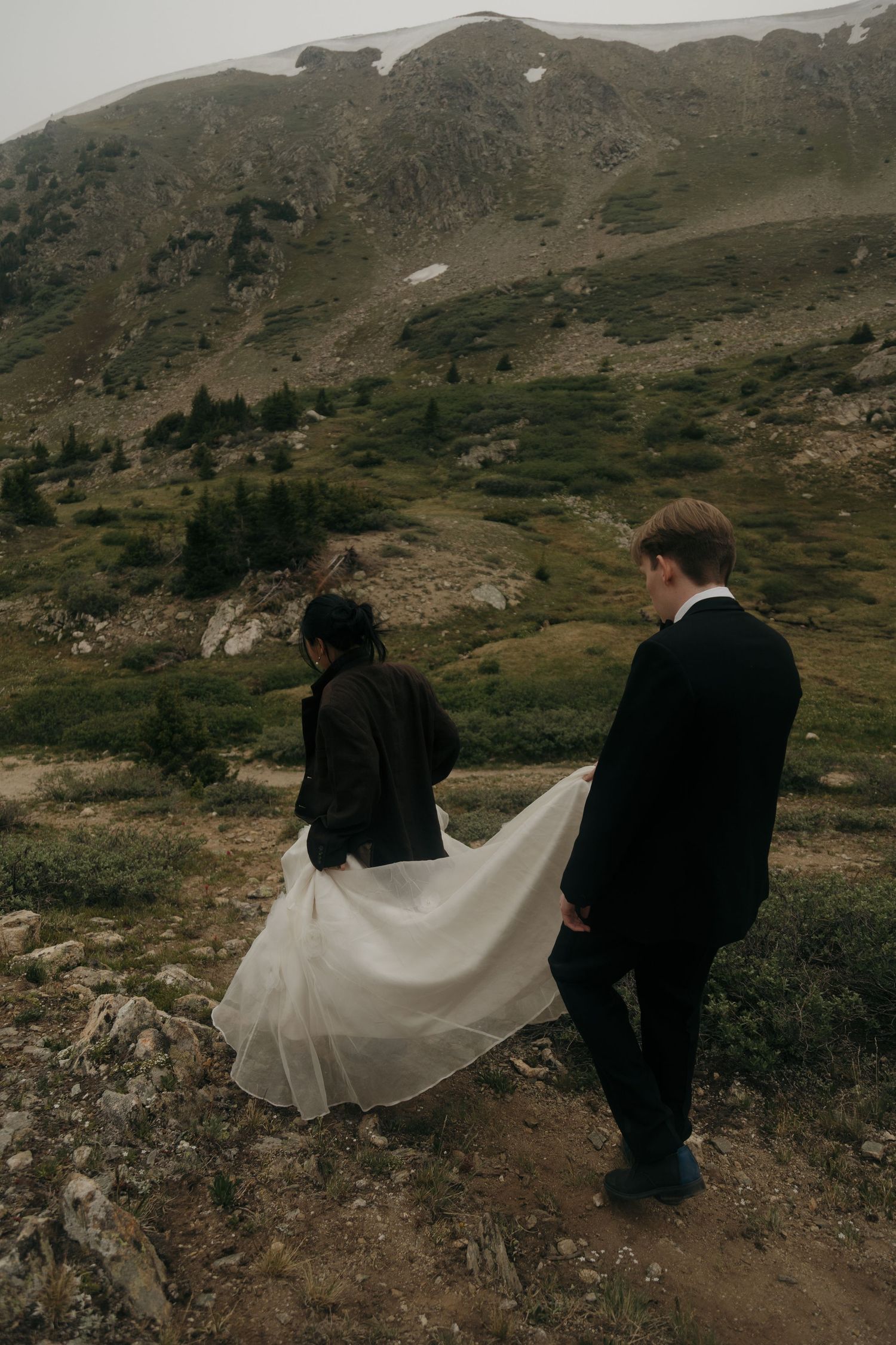 Bride and groom walk up a rocky mountain trail, her white gown flowing in the wind during their outdoor elopement.
