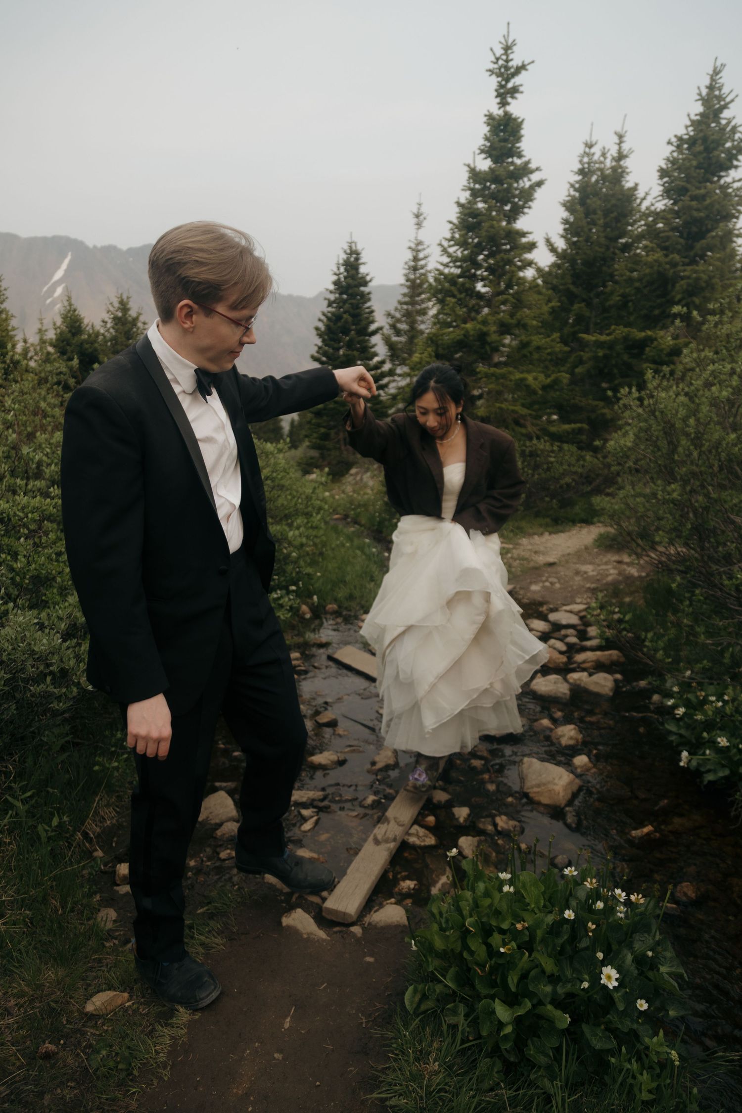 Groom helps bride cross a rocky stream on a mountain trail surrounded by pine trees.