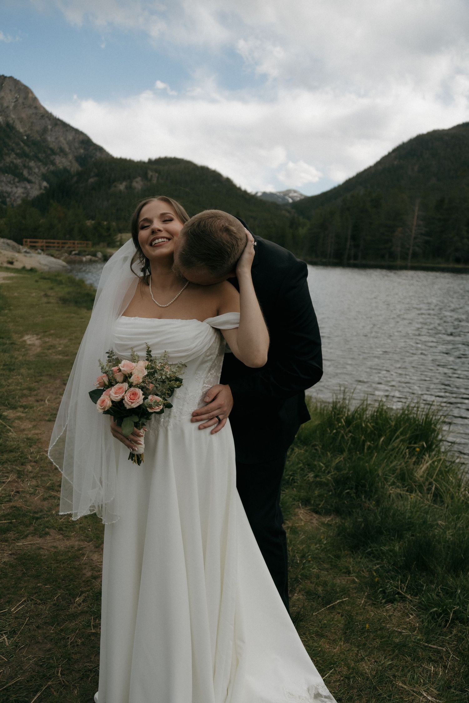 Bride laughing as groom kisses her cheek by a mountain lake, holding pink bouquet in white off-shoulder gown.