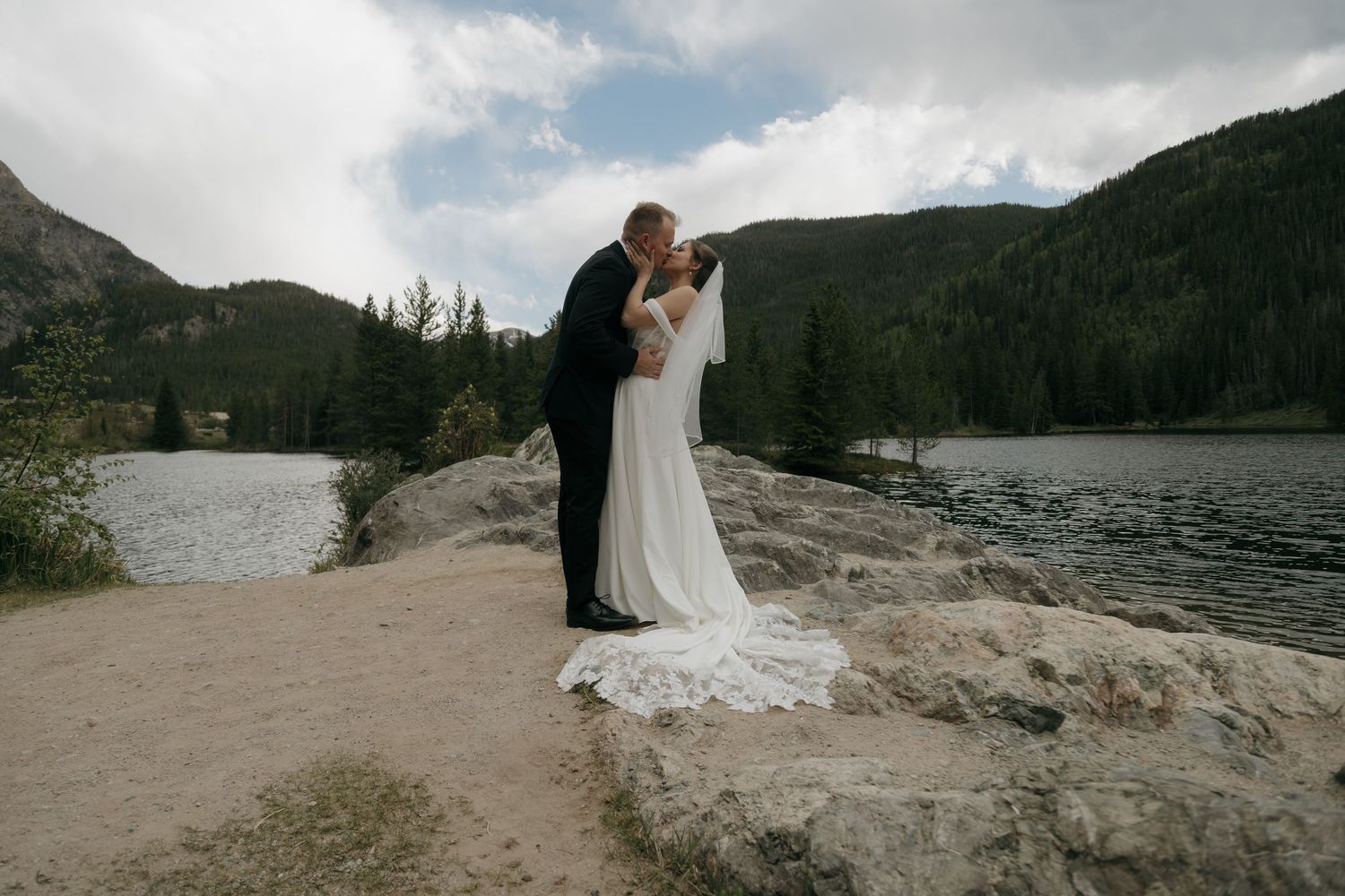 Bride and groom share a romantic kiss on rocky lakeside with mountain forest backdrop on their wedding day.