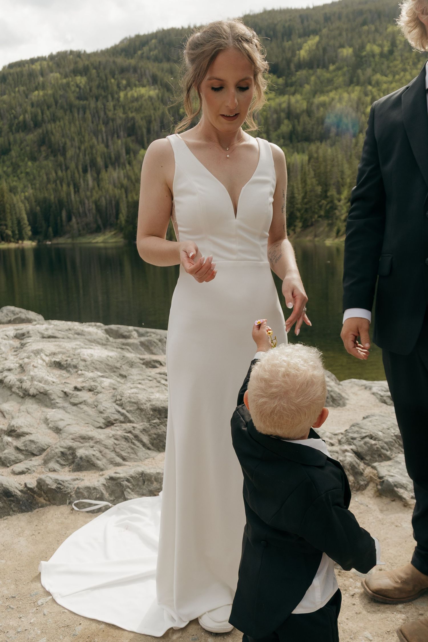 Bride in white gown interacts with young child in black suit by mountain lake.