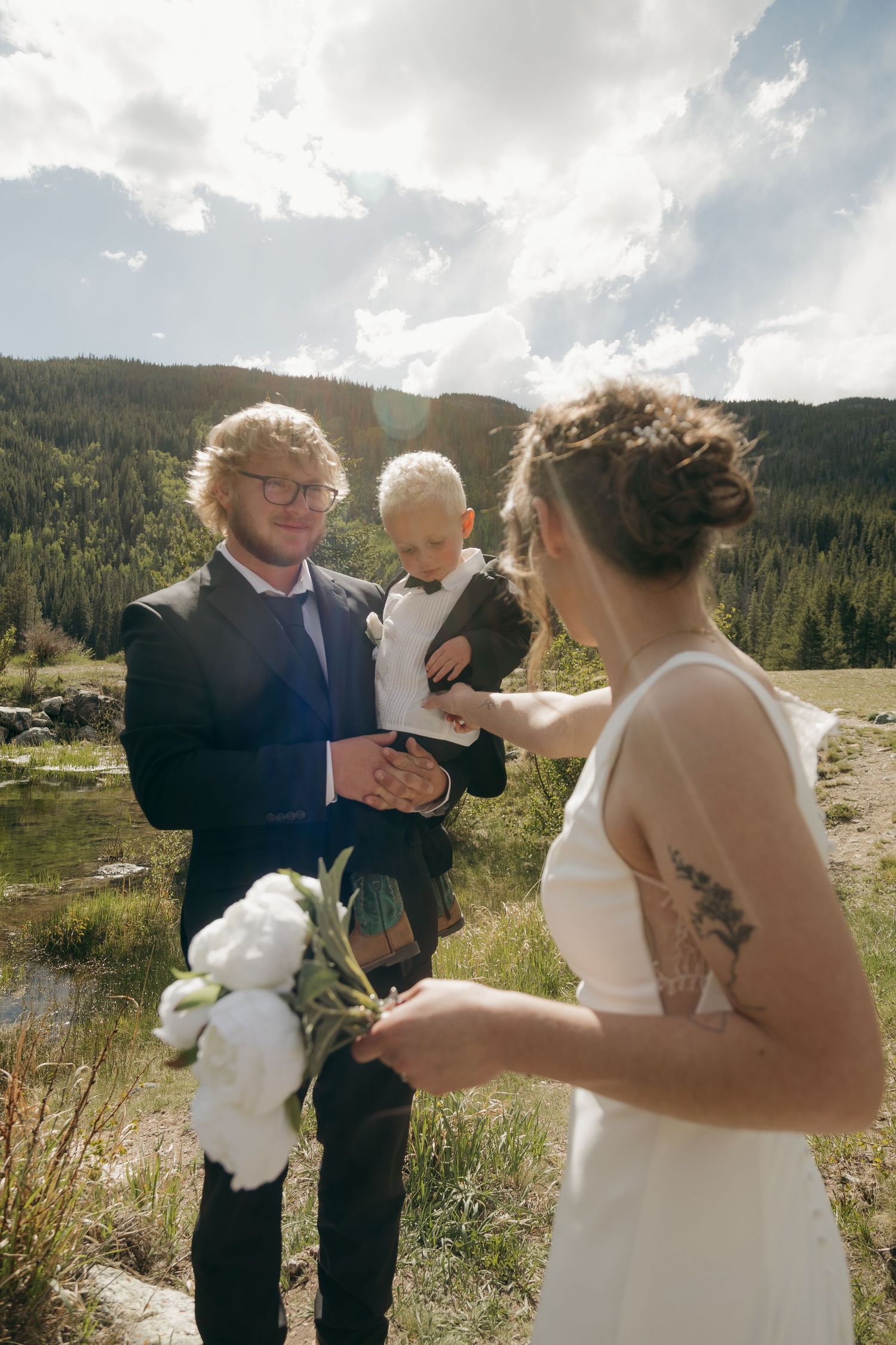 A groom holding a baby reaches toward his bride in a white dress during an outdoor mountain wedding ceremony.