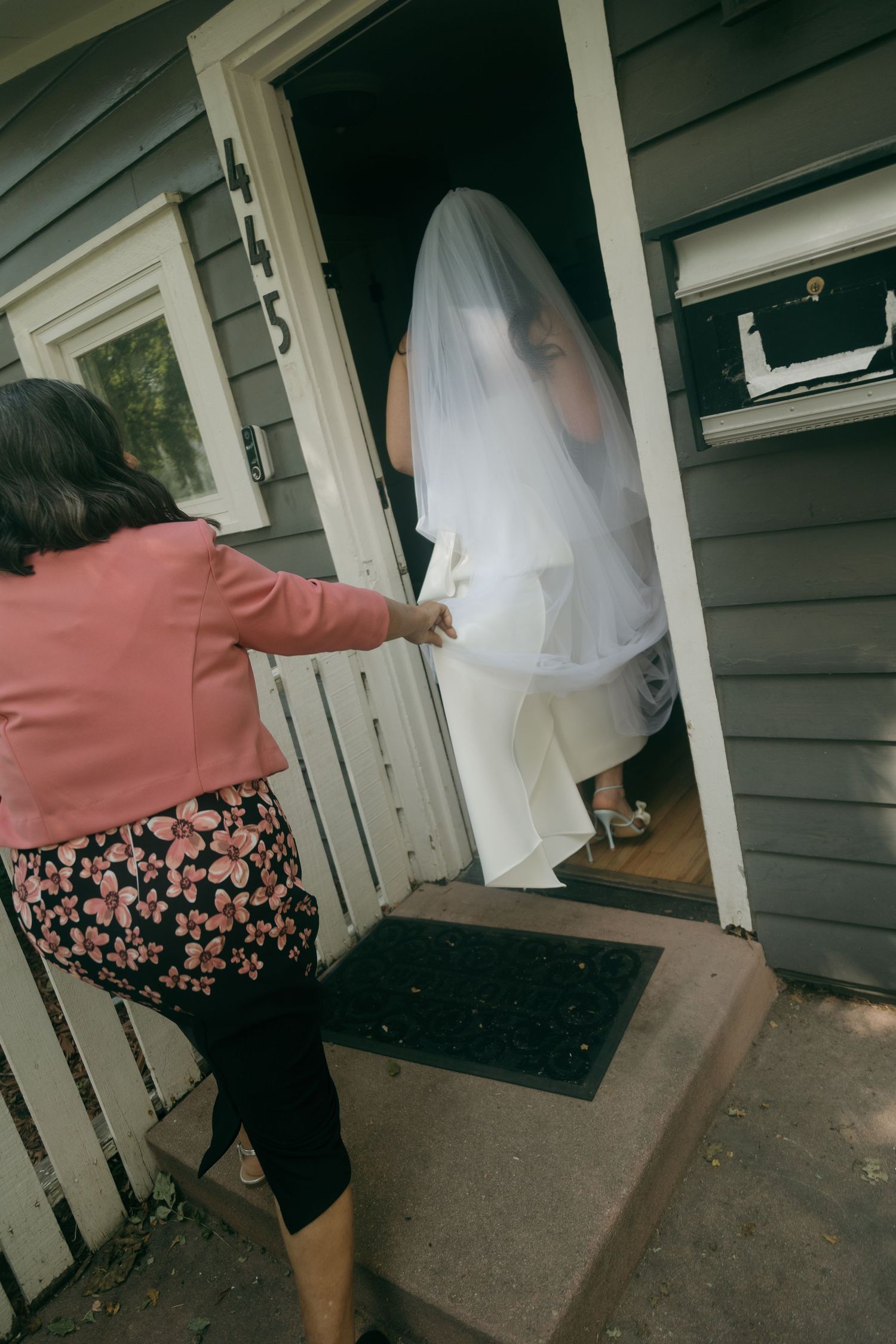 A bride in a white veil stands in a doorway as a woman in a pink jacket reaches toward her on the front steps.