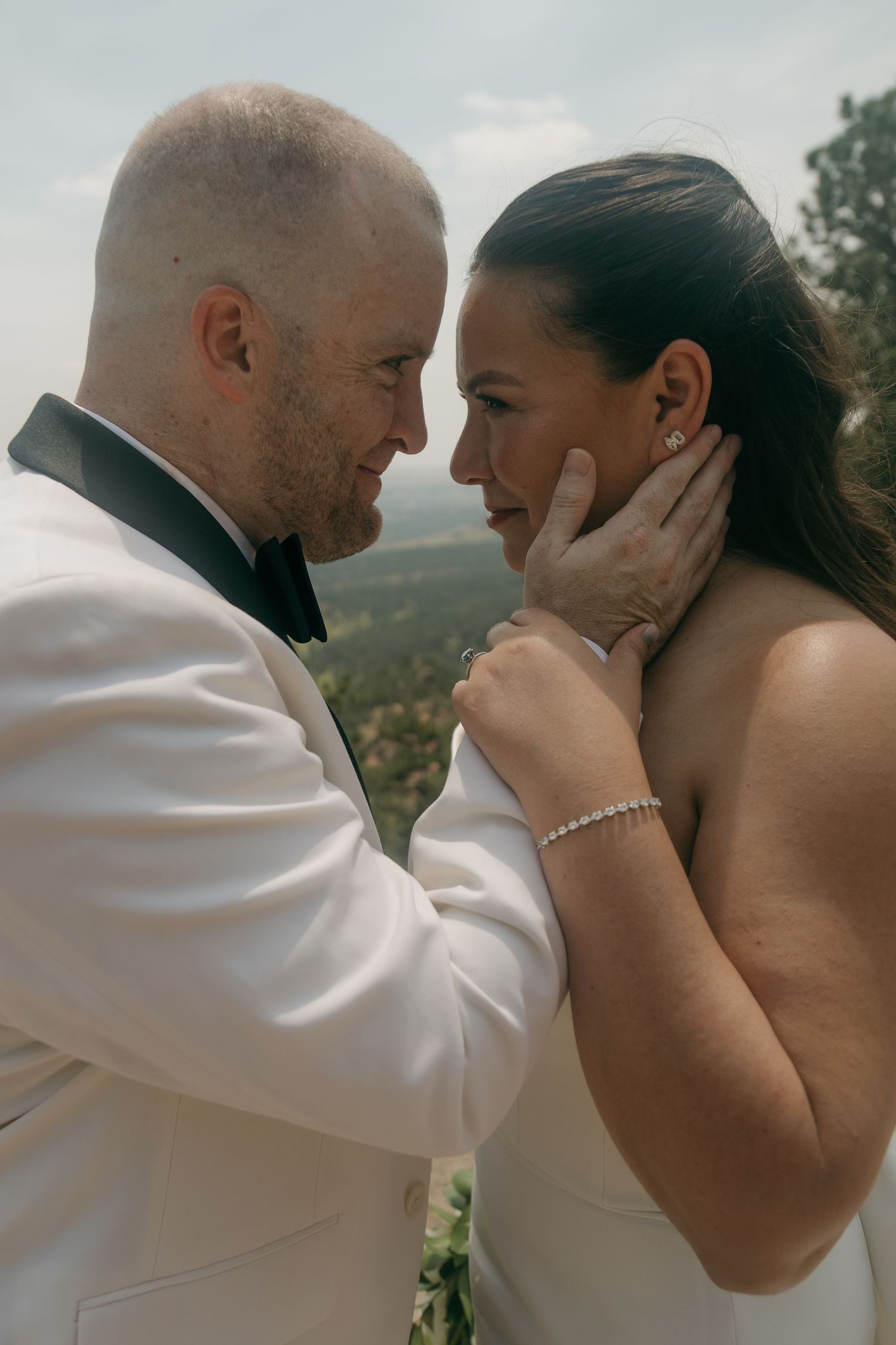 A groom in a white tuxedo tenderly cups the face of his bride with a scenic mountain backdrop behind them.