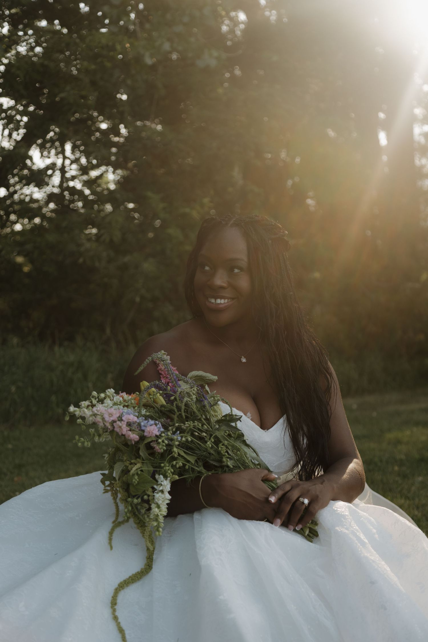 Smiling Black bride in white ball gown holding wildflower bouquet outdoors in golden sunset light.