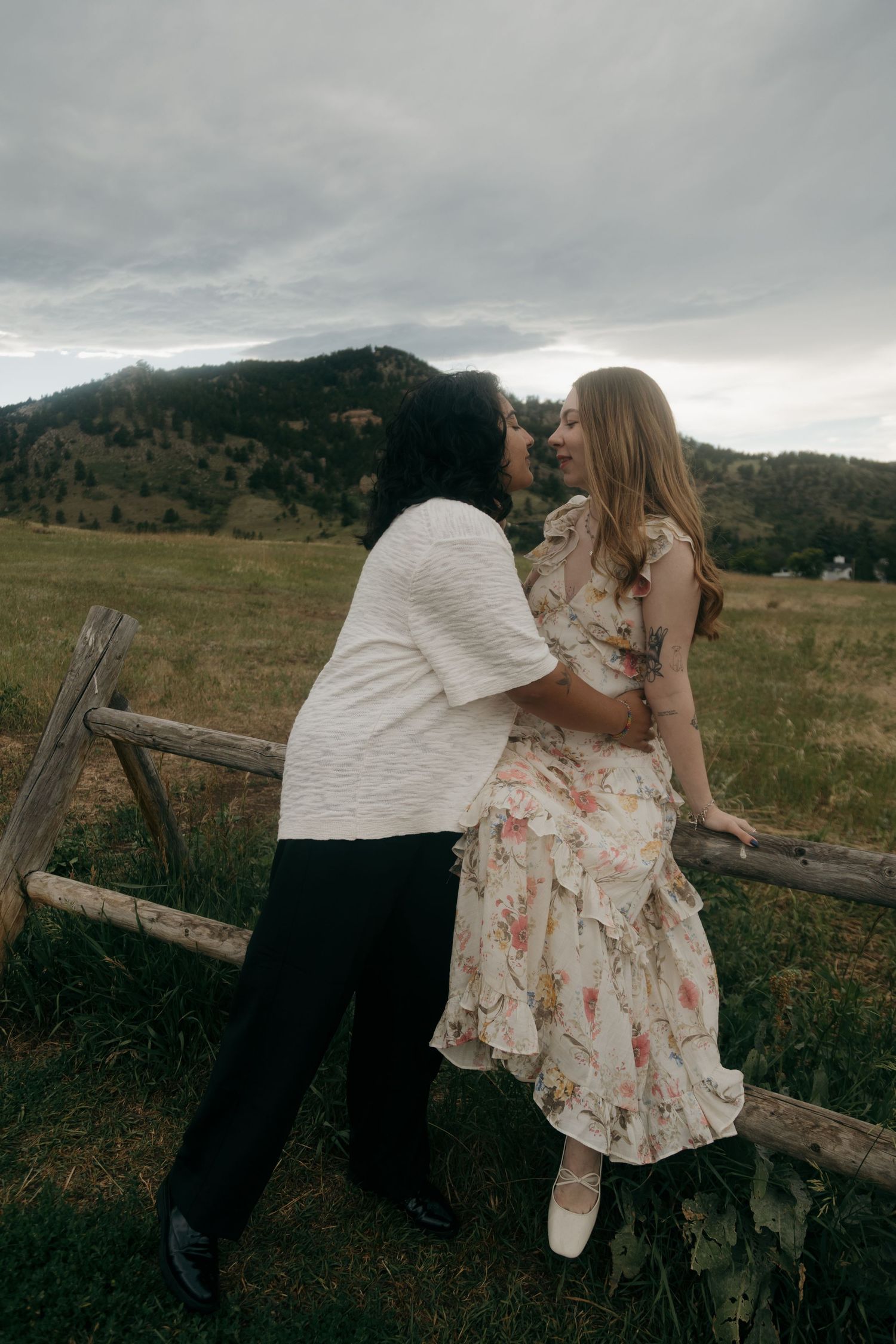 Couple sharing a kiss near a rustic wooden fence with rolling green hills behind them.