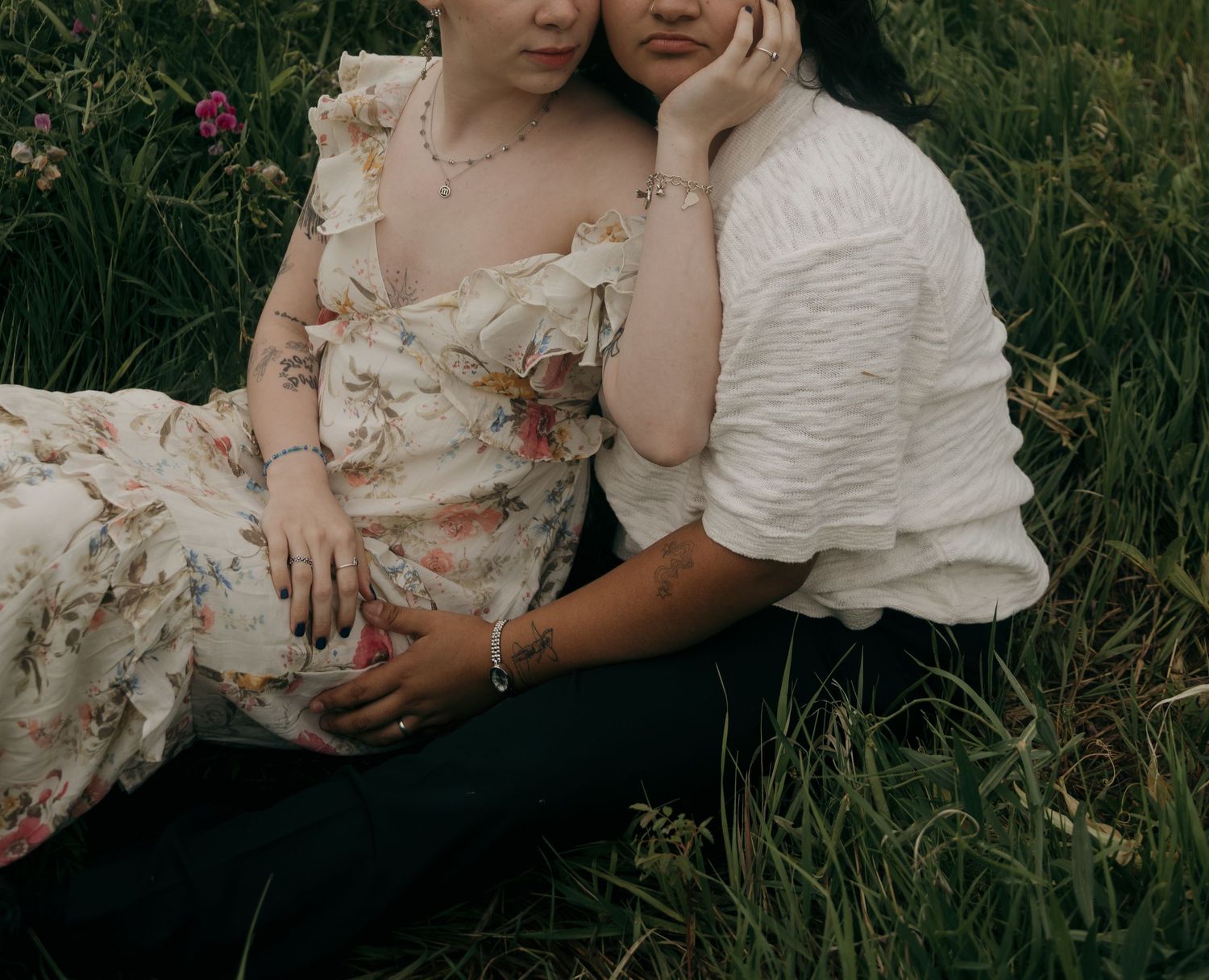 Couple sitting in green grass, woman in floral dress leaning against partner in white textured shirt.