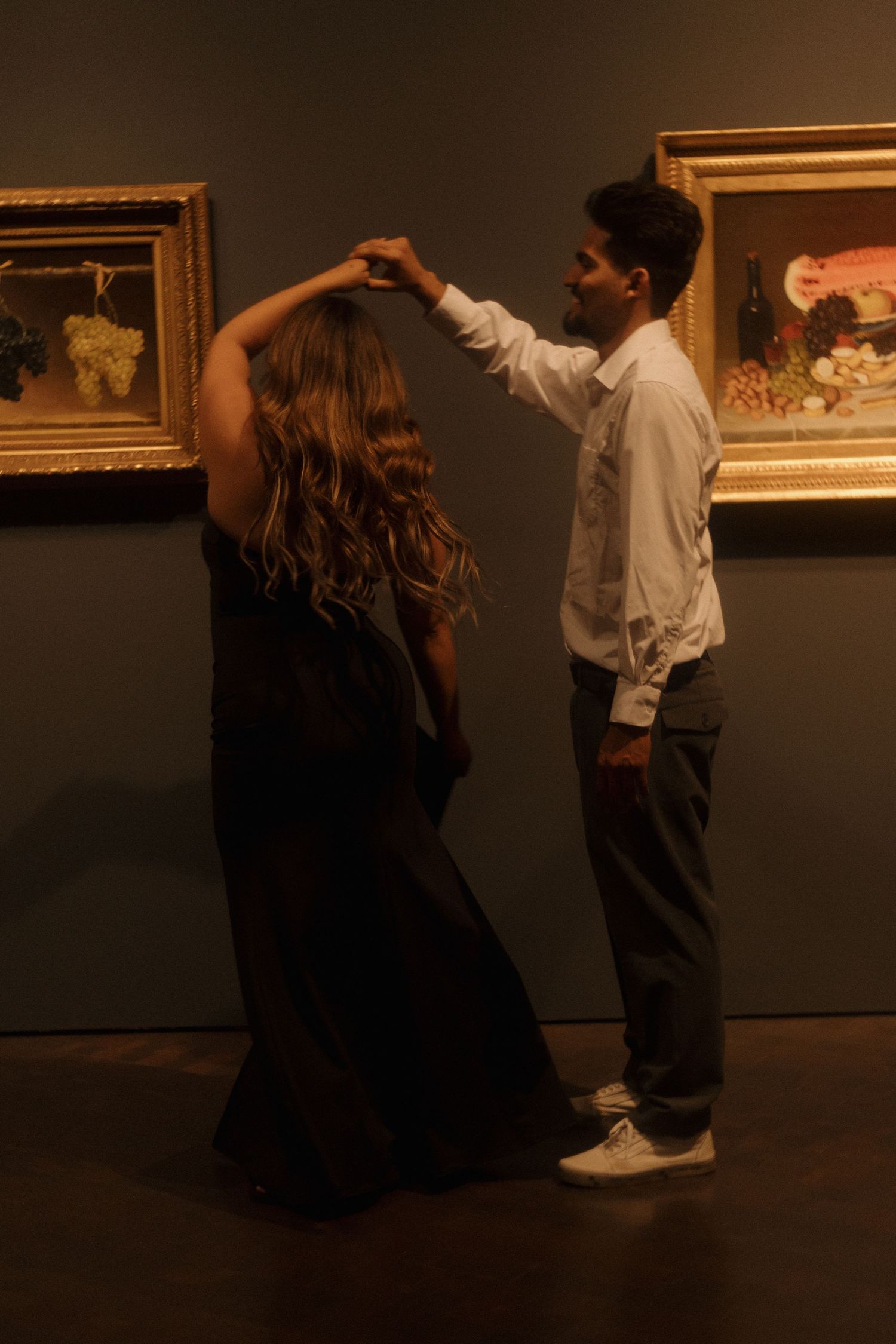 Couple dancing in a dimly lit art museum, man spinning woman in black dress near golden-framed paintings.
