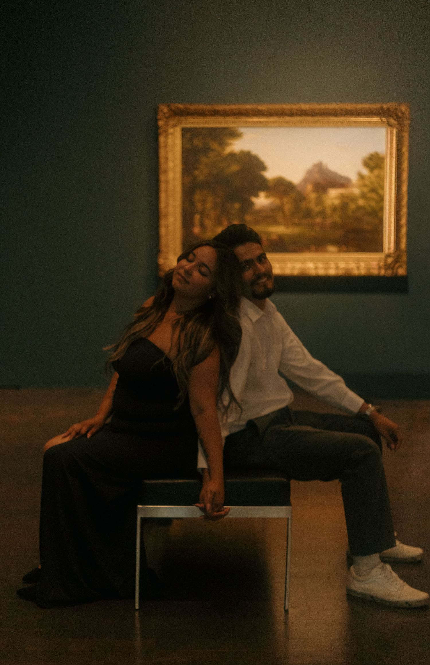 Couple sitting back-to-back on a bench in a dimly lit art museum gallery.