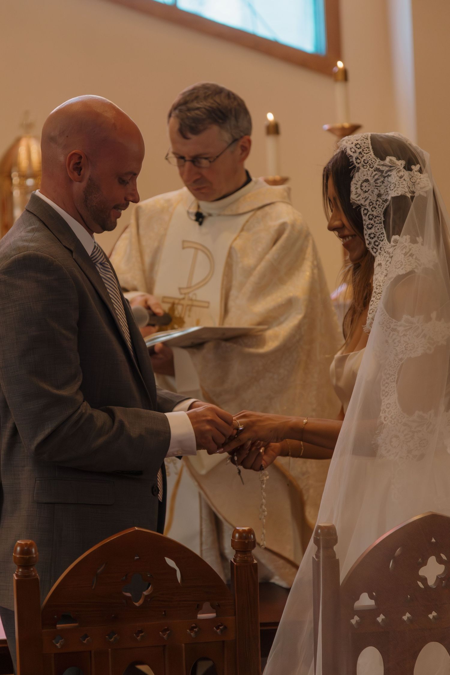 Groom places wedding ring on bride's finger as priest officiates Catholic ceremony.